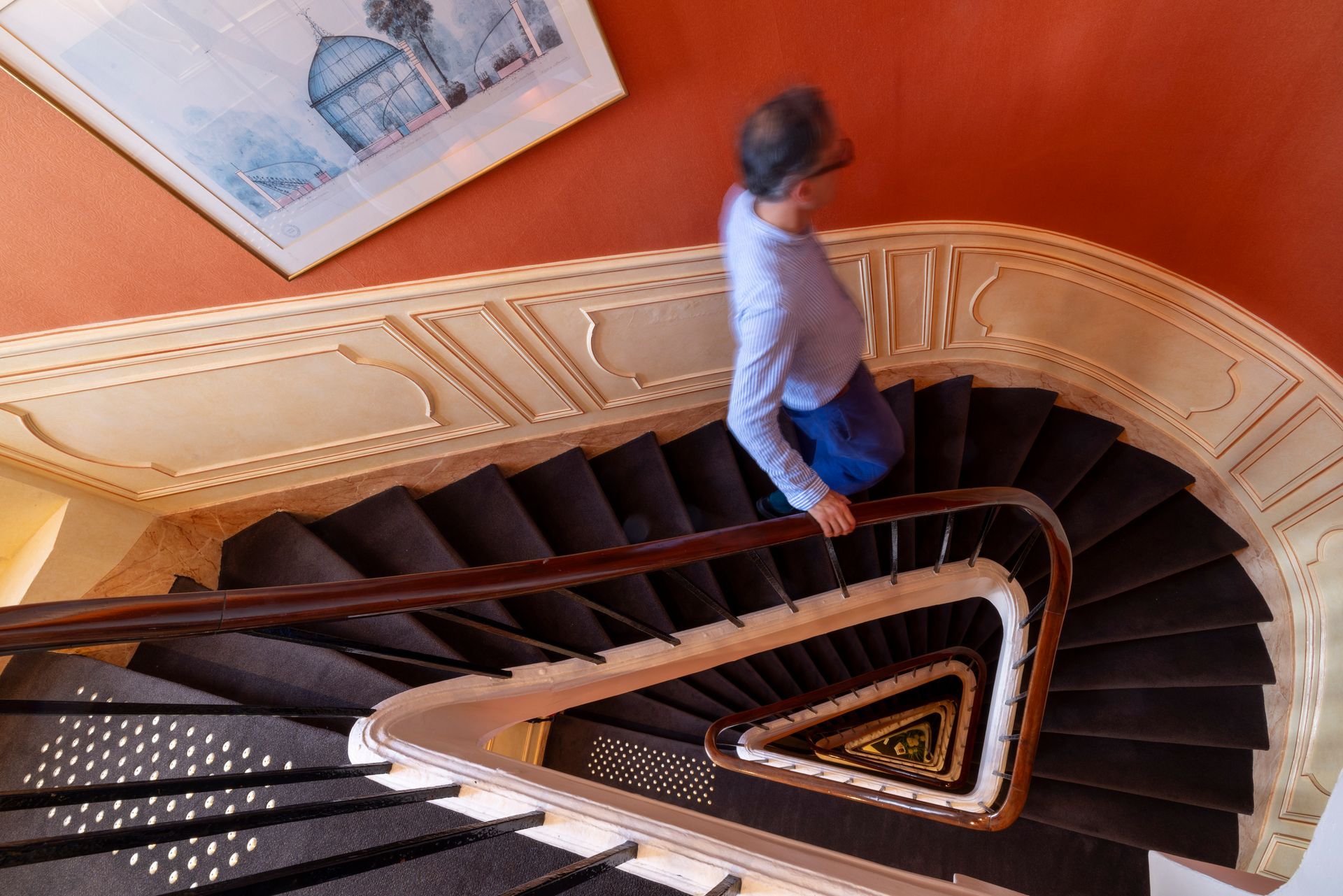 Personne descendant un escalier sombre en colimaçon avec une rampe en bois. Mur orange, œuvre d'art encadrée.