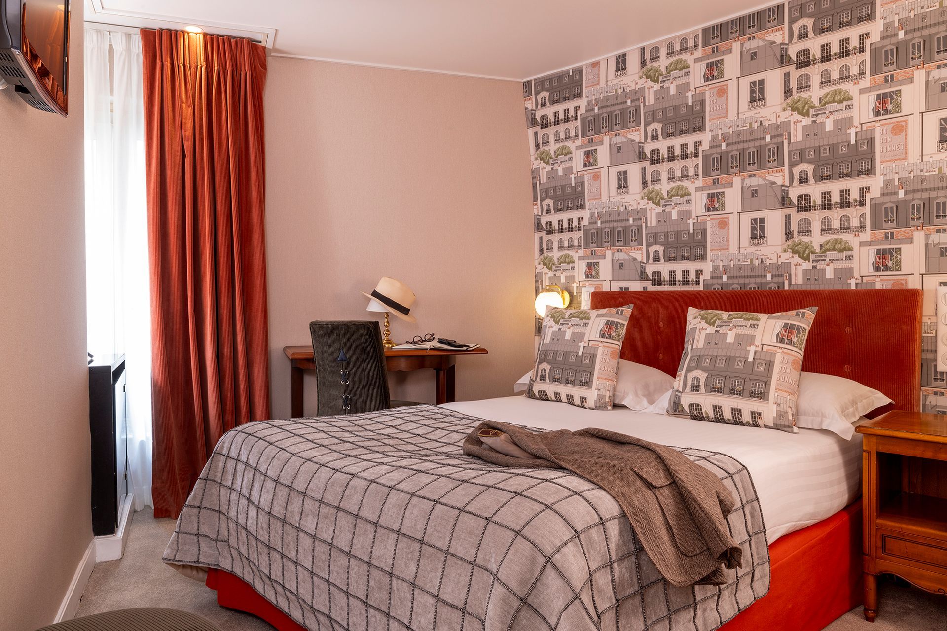 Chambre d'hôtel avec lit, bureau et papier peint à motifs urbains. Rideaux, cadre de lit et jeté de lit orange.