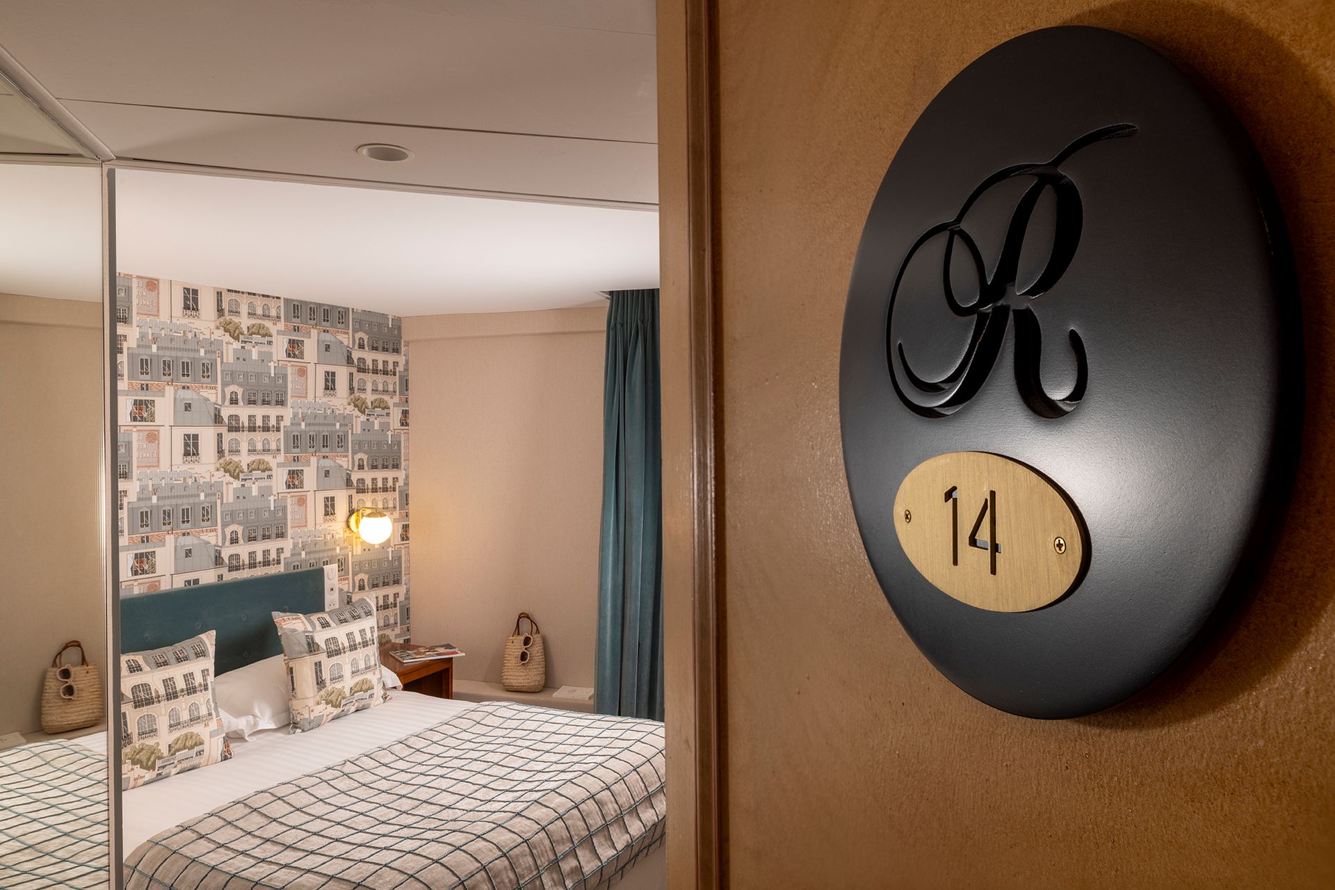 La porte d'une chambre d'hôtel portant le numéro 14. Derrière la porte ouverte se trouve un lit orné de coussins décoratifs.