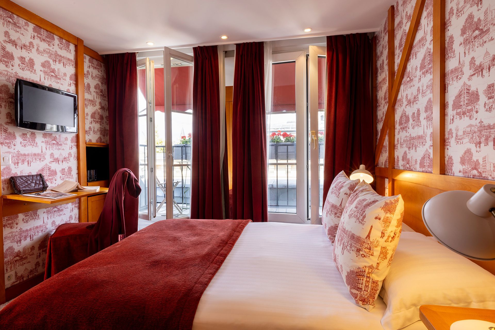 Chambre d'hôtel avec grand lit, balcon, papier peint, fleuri rouge et blanc, bureau et télévision.