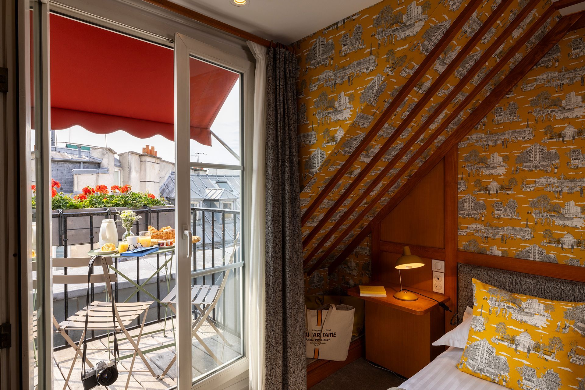 Chambre d'hôtel avec balcon : papier peint à motifs jaunes, porte ouverte, store rouge, table dressée pour le petit-déjeuner.