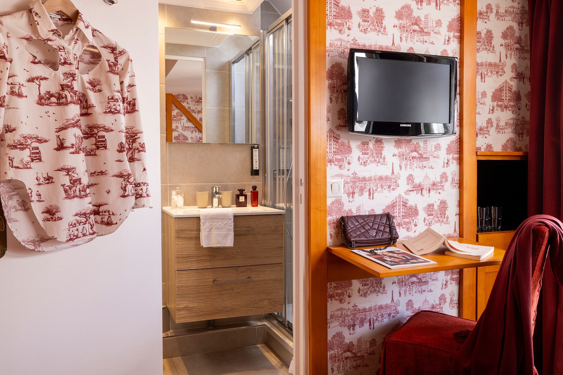 Chambre d'hôtel avec coiffeuse, douche, télévision, petit bureau, papier peint à motifs rouges et une chemise accrochée à la porte.