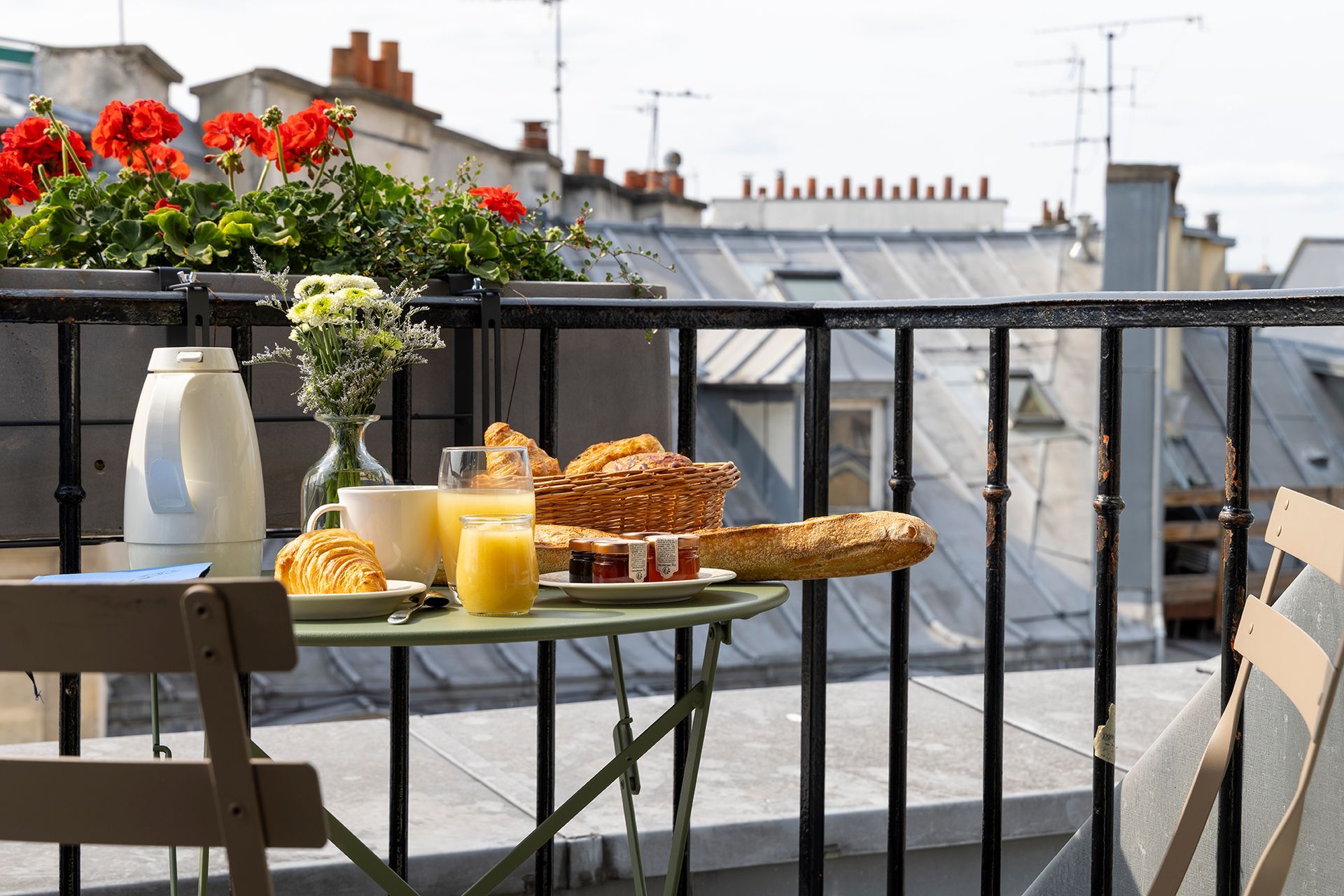 Petit-déjeuner sur un balcon parisien : table dressée avec nourriture et boissons, fleurs rouges en arrière-plan, toits de la ville.