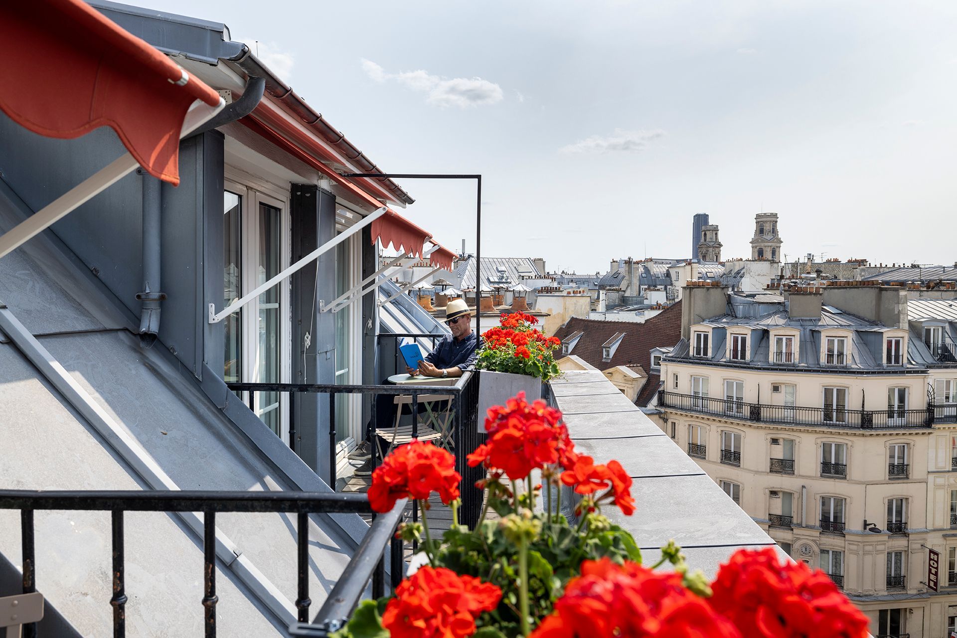 Balcon orné de fleurs rouges donnant sur les toits parisiens, avec une personne lisant à une table.