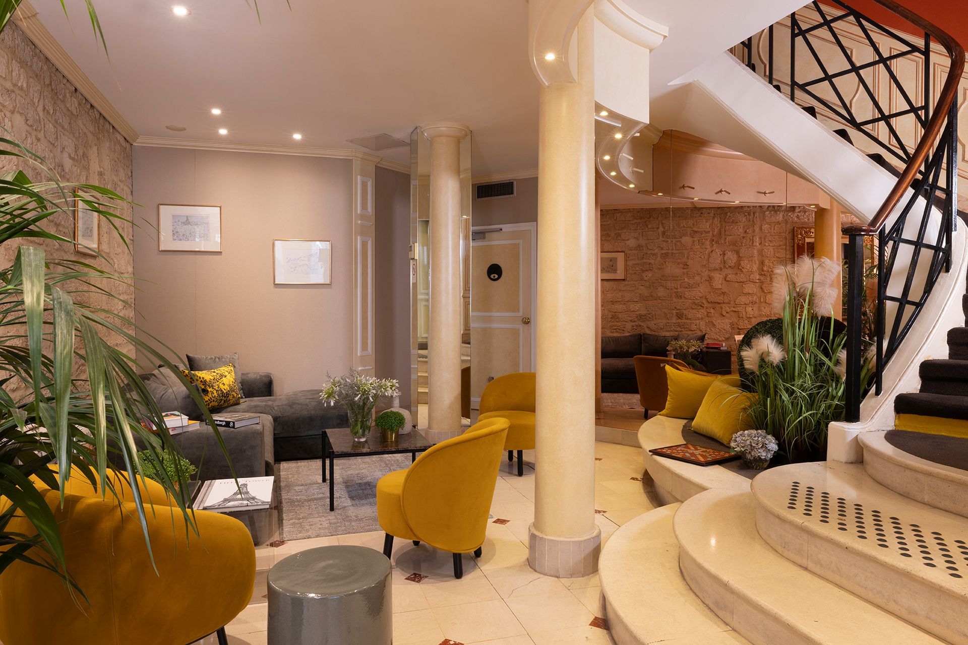 Hall d'entrée avec fauteuils jaunes, canapé gris, escalier en colimaçon et plantes. Murs en pierre et couleur crème.