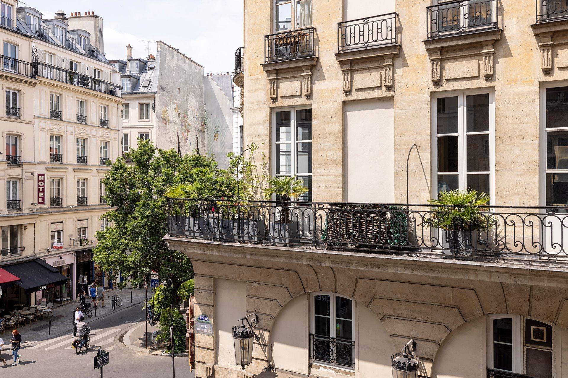 Coin de rue parisien avec balcons, balustrades ouvragées et immeubles. On aperçoit des passants et des véhicules.