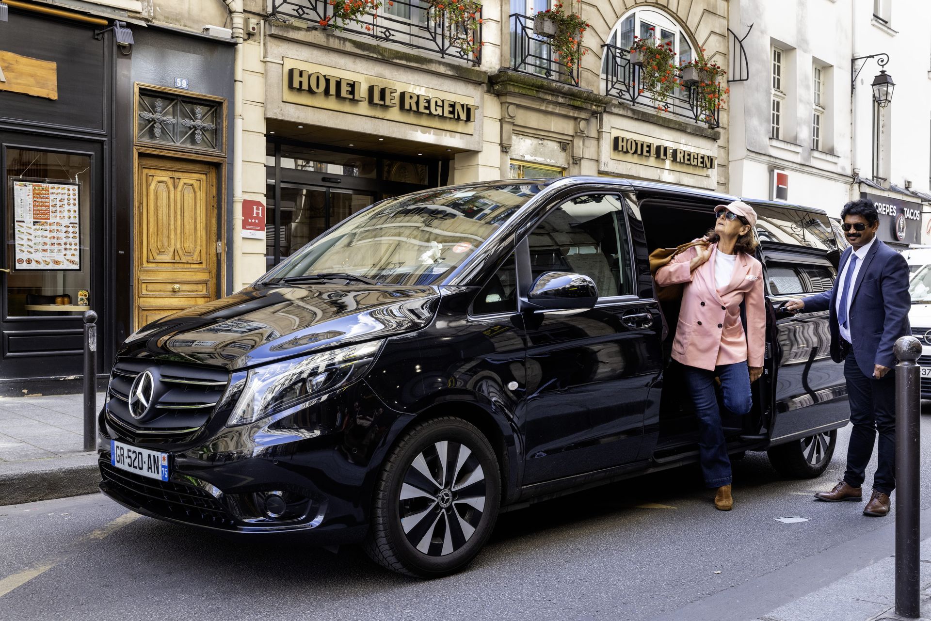 Une camionnette Mercedes noire est garée devant un bâtiment. Une personne descend du véhicule tandis qu'une autre se tient près de la portière.