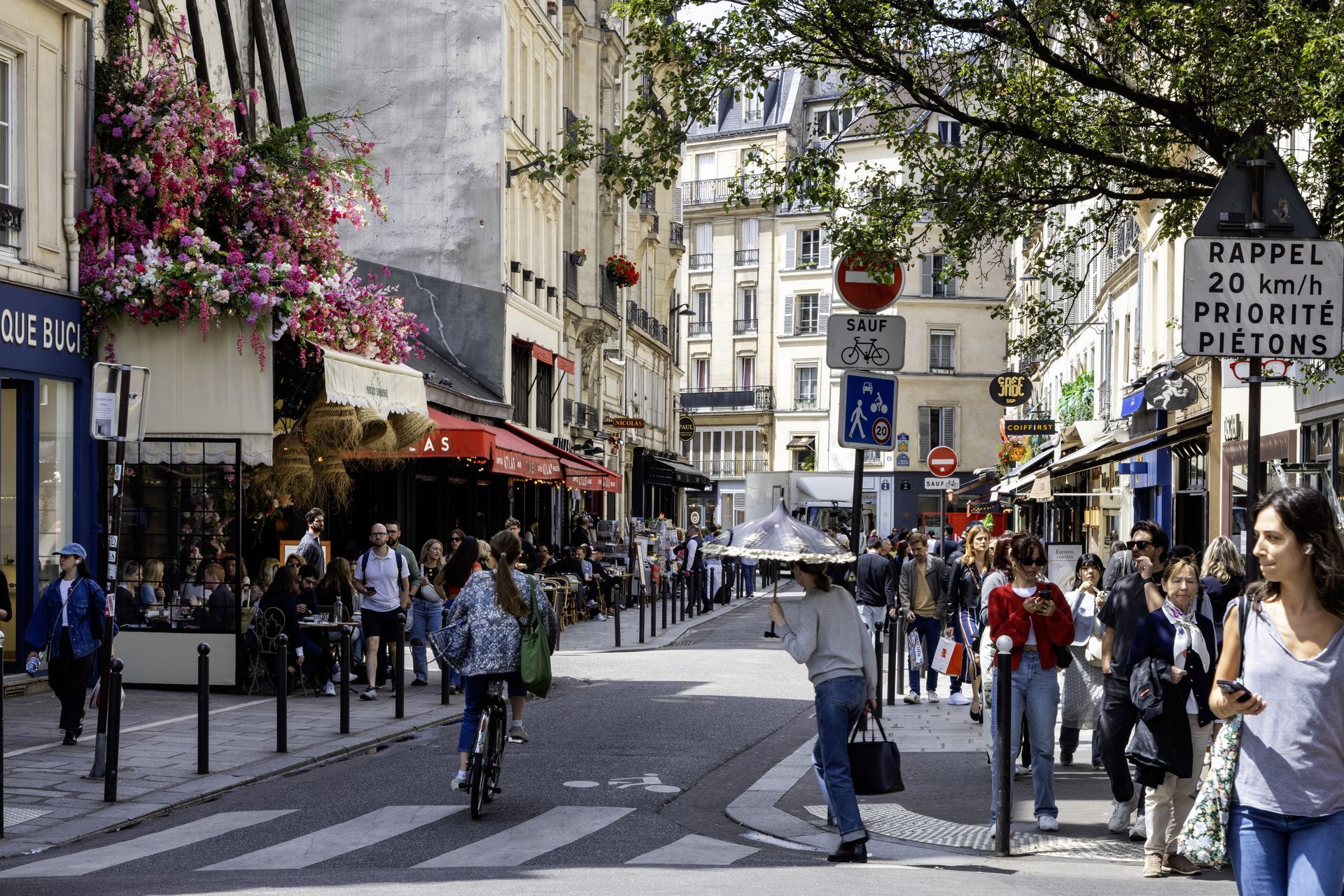 Scène de rue animée à Paris : des passants, des boutiques, des restaurants avec terrasses, un cycliste et des panneaux de signalisation.