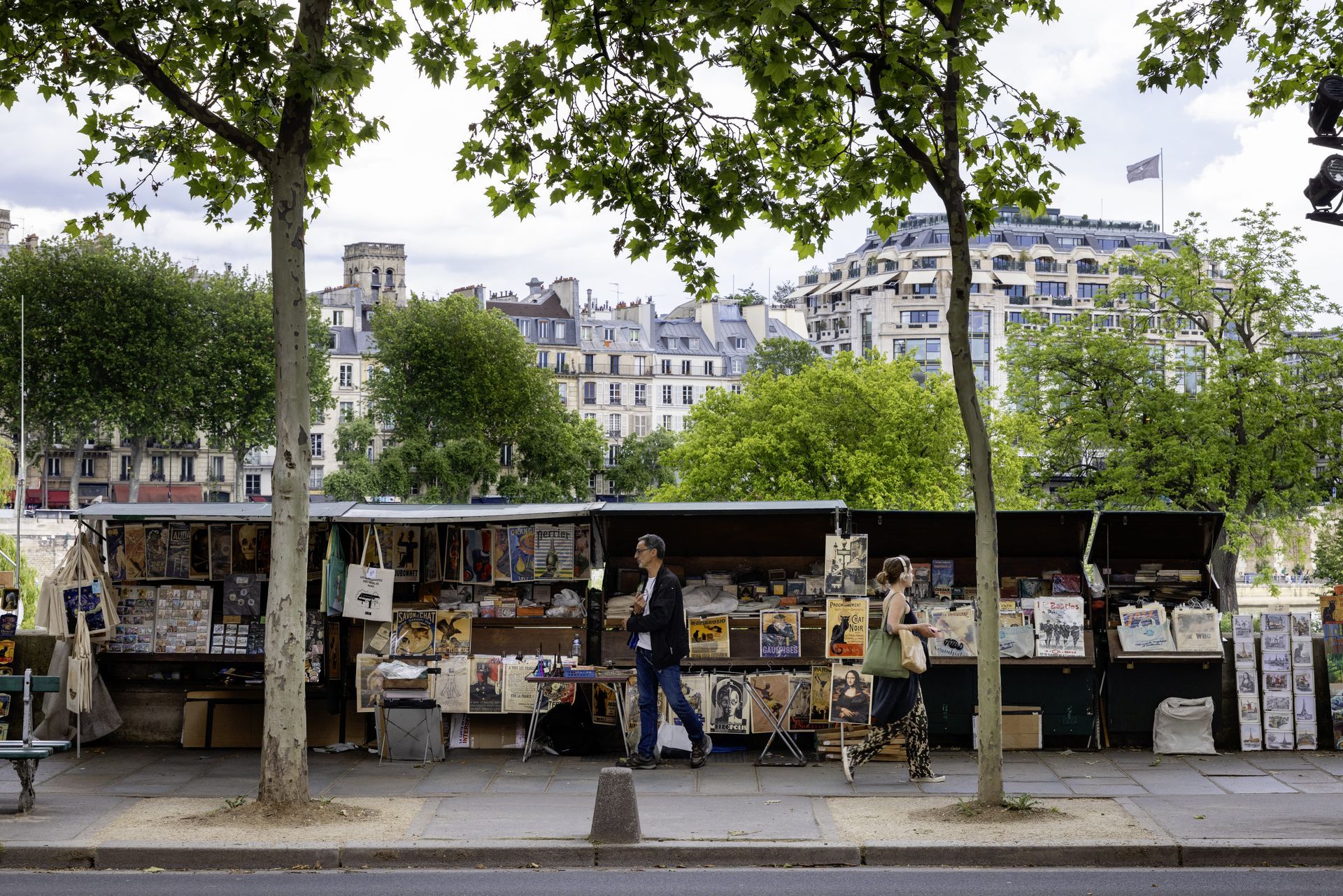 Des étals de livres bordent une rue parisienne, deux personnes passent devant. Des immeubles et des arbres se dessinent à l'arrière-plan.