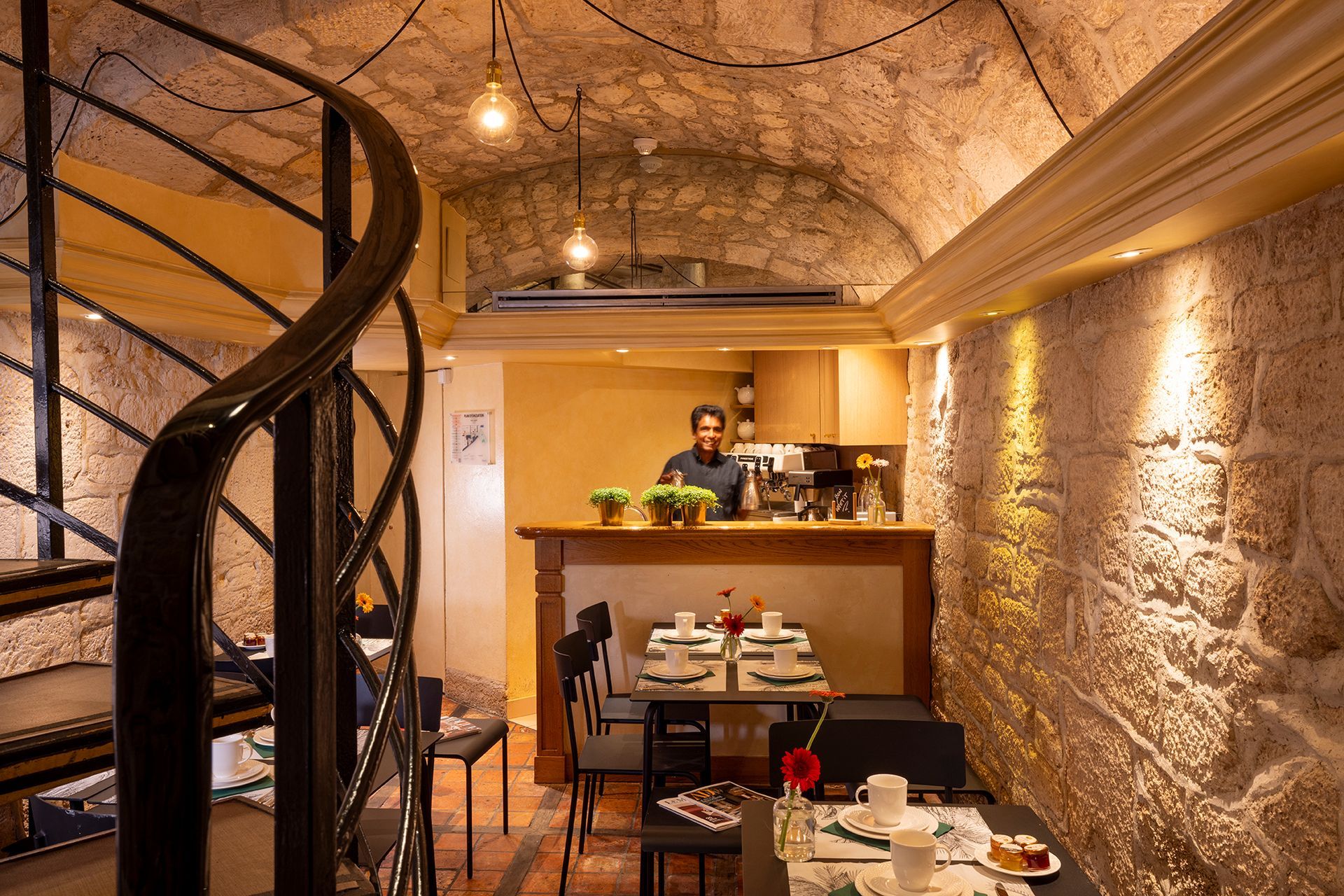 Intérieur d'un petit café avec des murs en pierre, un escalier en colimaçon et une personne derrière le comptoir.