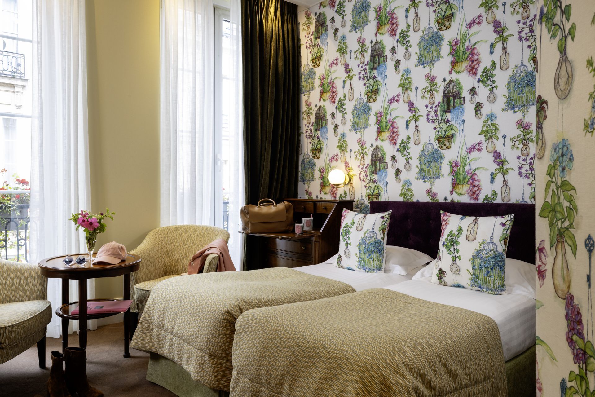 Deux lits jumeaux dans une chambre d'hôtel avec papier peint à fleurs, une petite table, des chaises et une fenêtre.