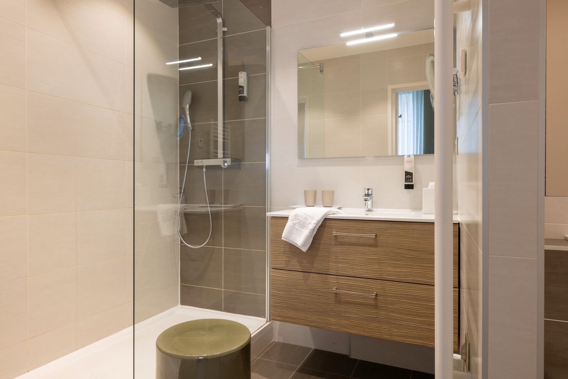 Salle de bains avec douche en verre, meuble-lavabo avec armoire en bois, miroir et tabouret.