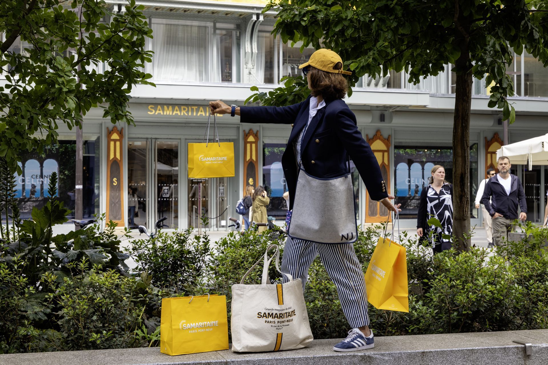 Une personne portant des sacs de courses devant un magasin. Sacs jaune et blanc ; un bâtiment aux ornements dorés se dresse en arrière-plan.