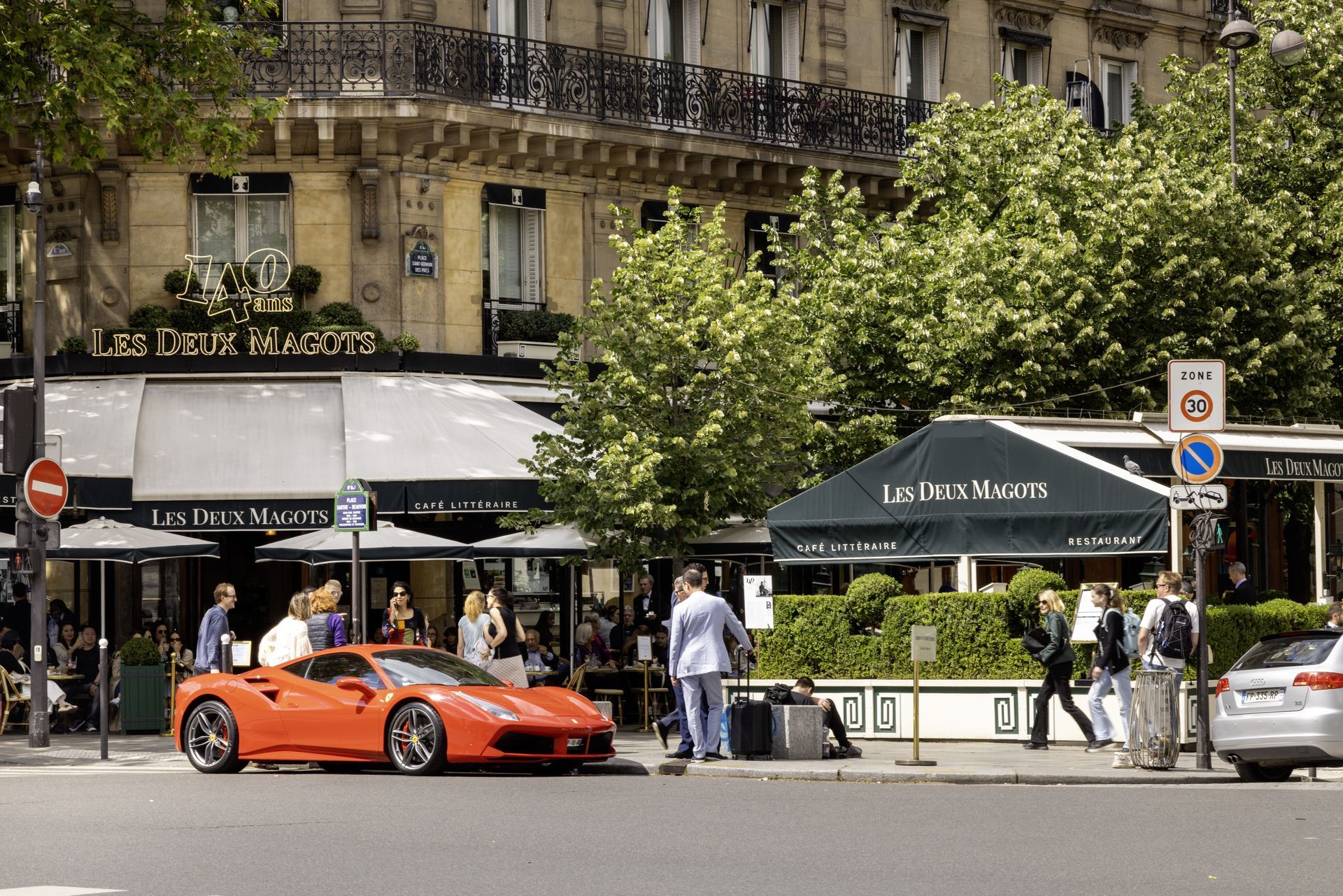 Ferrari orange garée dans la rue près d'un café, des clients déjeunent en terrasse. Paris, France.