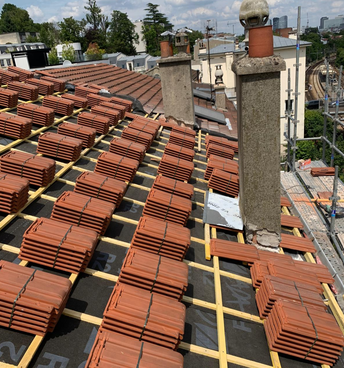 Toiture en construction avec tuiles rouges empilées, poutres apparentes et deux cheminées en briques dans un quartier urbain