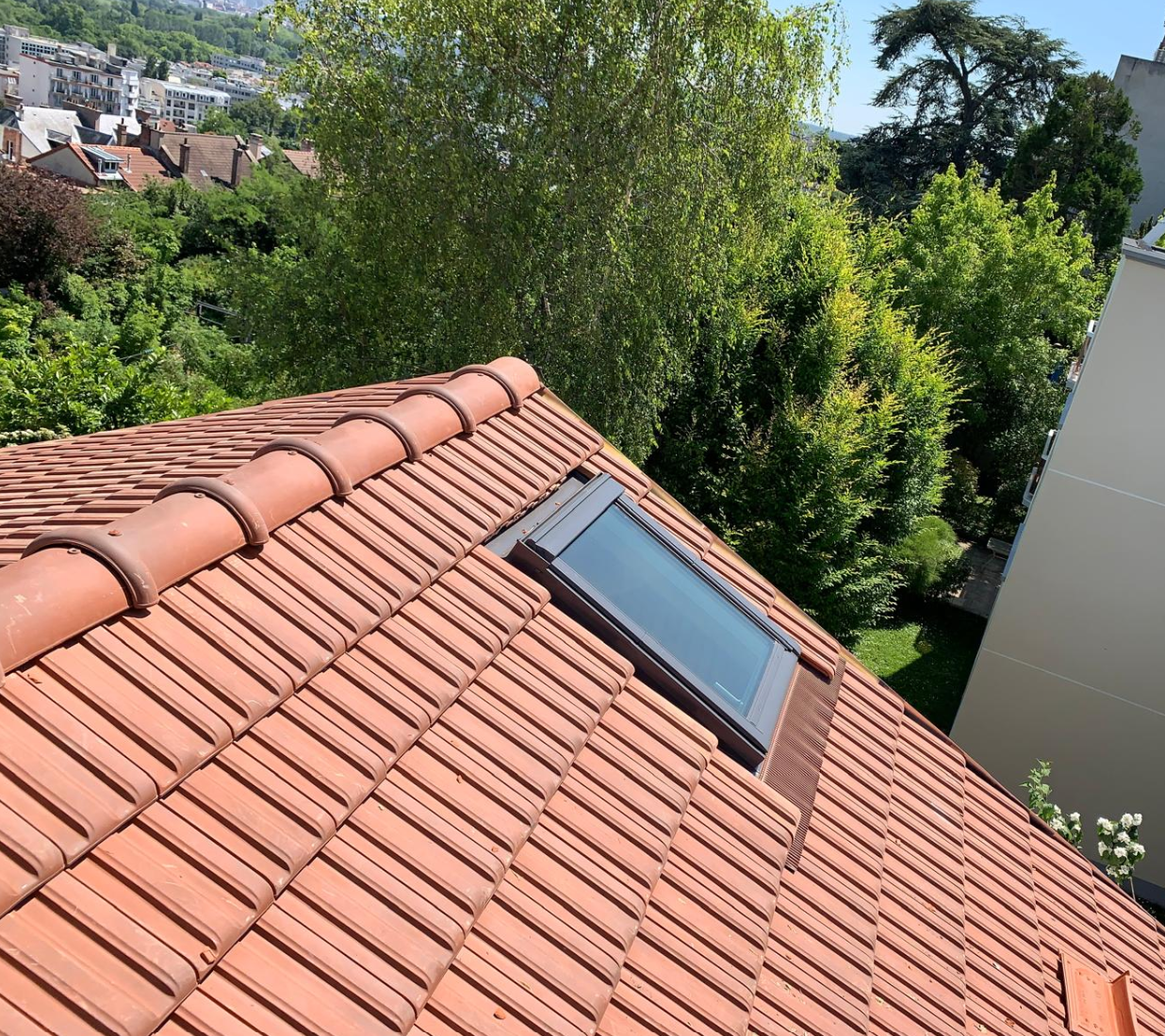 Toit en pente recouvert de tuiles en terre cuite avec un puits de lumière, donnant sur des arbres verdoyants et les maisons voisines.