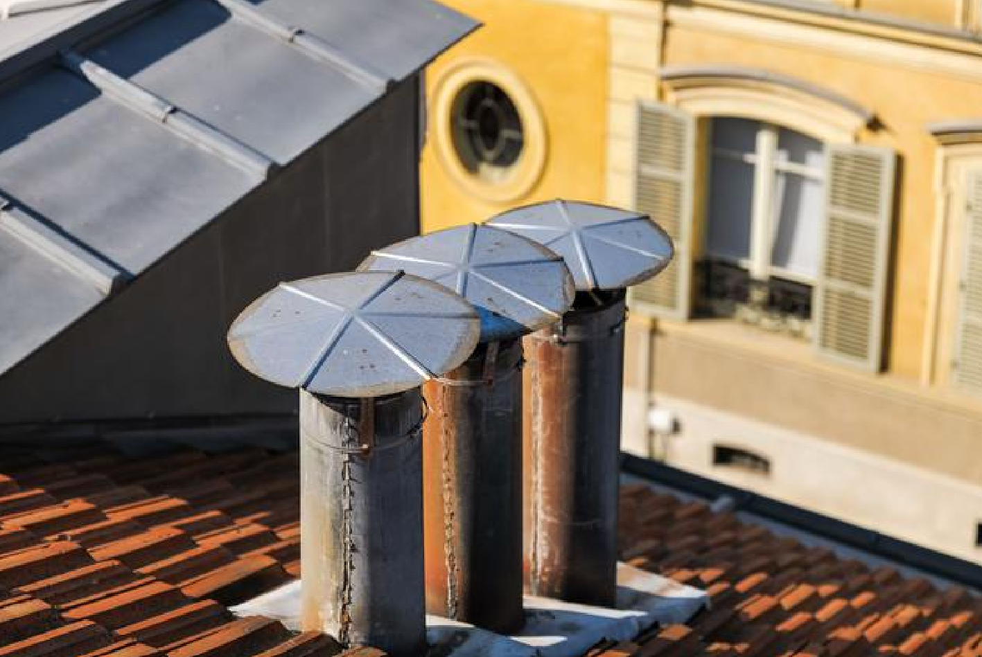 Trois grilles d'aération métalliques sur un toit de tuiles, avec un bâtiment en arrière-plan.
