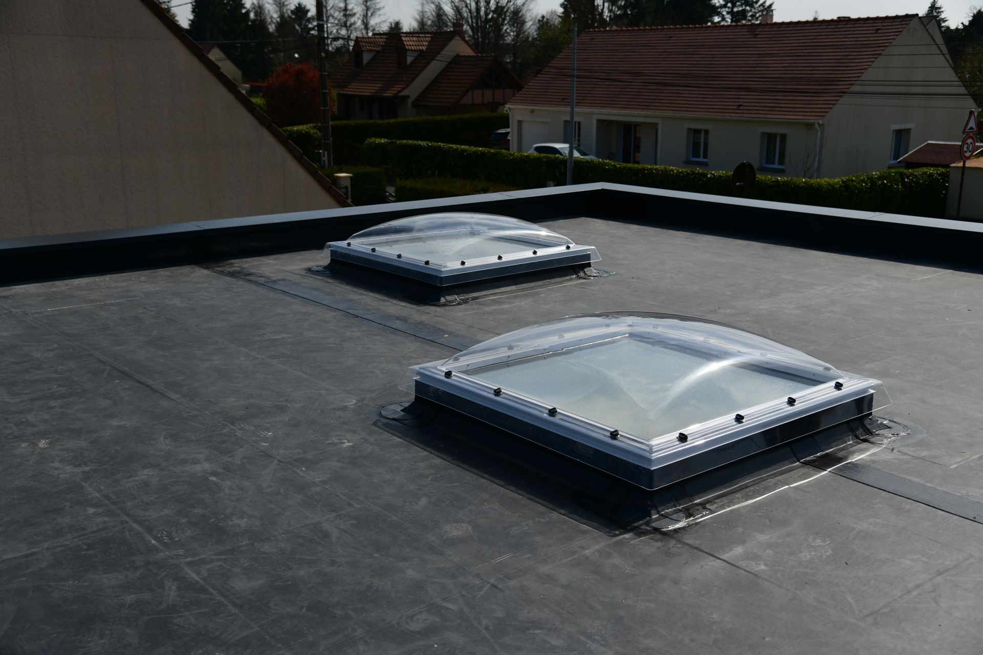 Toiture avec deux puits de lumière carrés sur un toit plat noir, avec des arbres et des maisons en arrière-plan