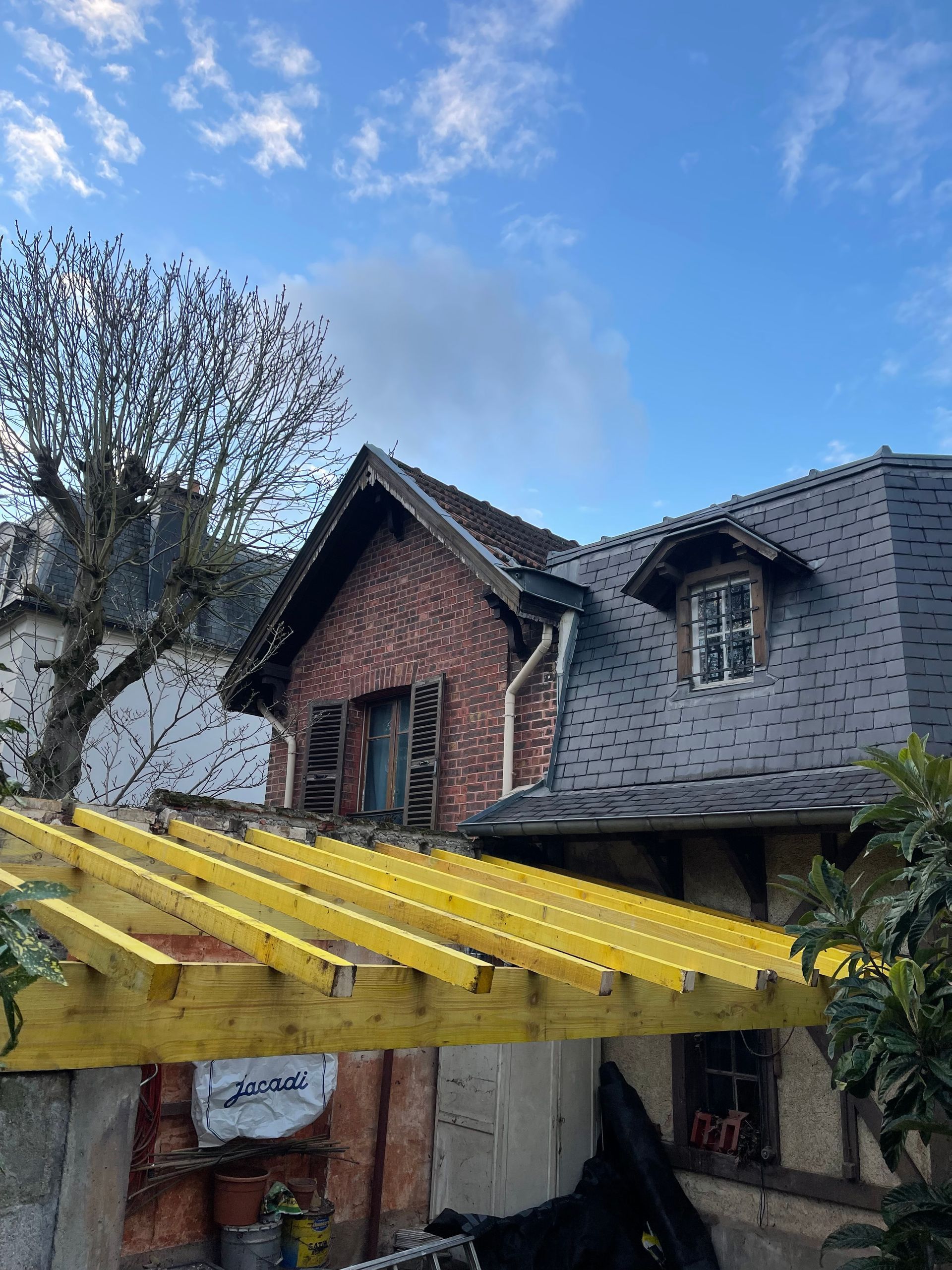 Pergola en bois jaune à côté d'une maison en briques au toit de bardeaux gris, sous un ciel bleu