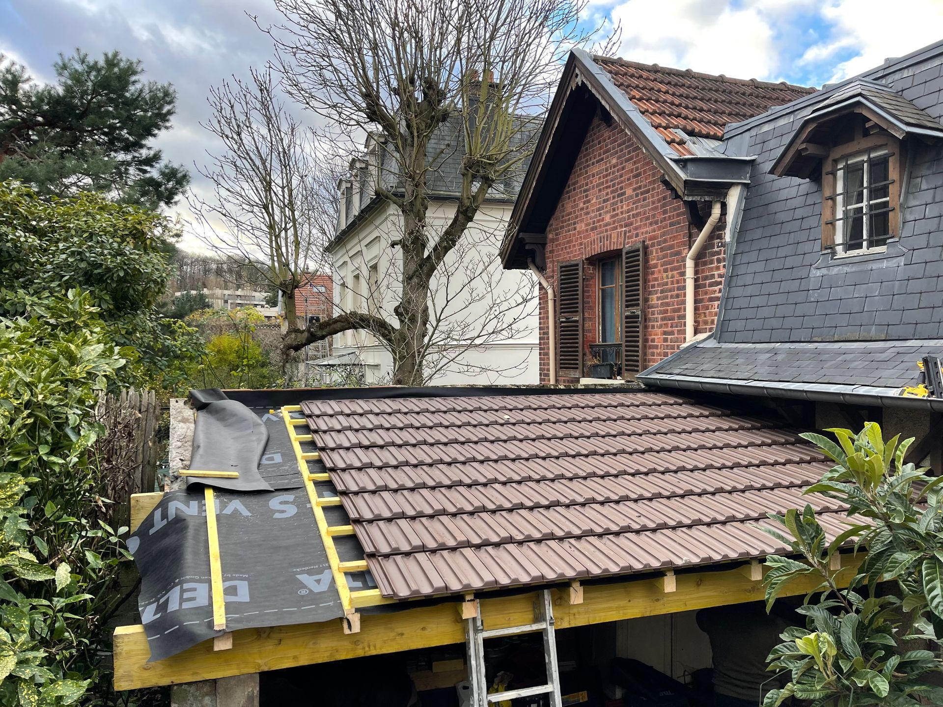 Réparation de toiture avec sous-couche noire et échelles jaunes à côté d'une maison en briques et d'arbres