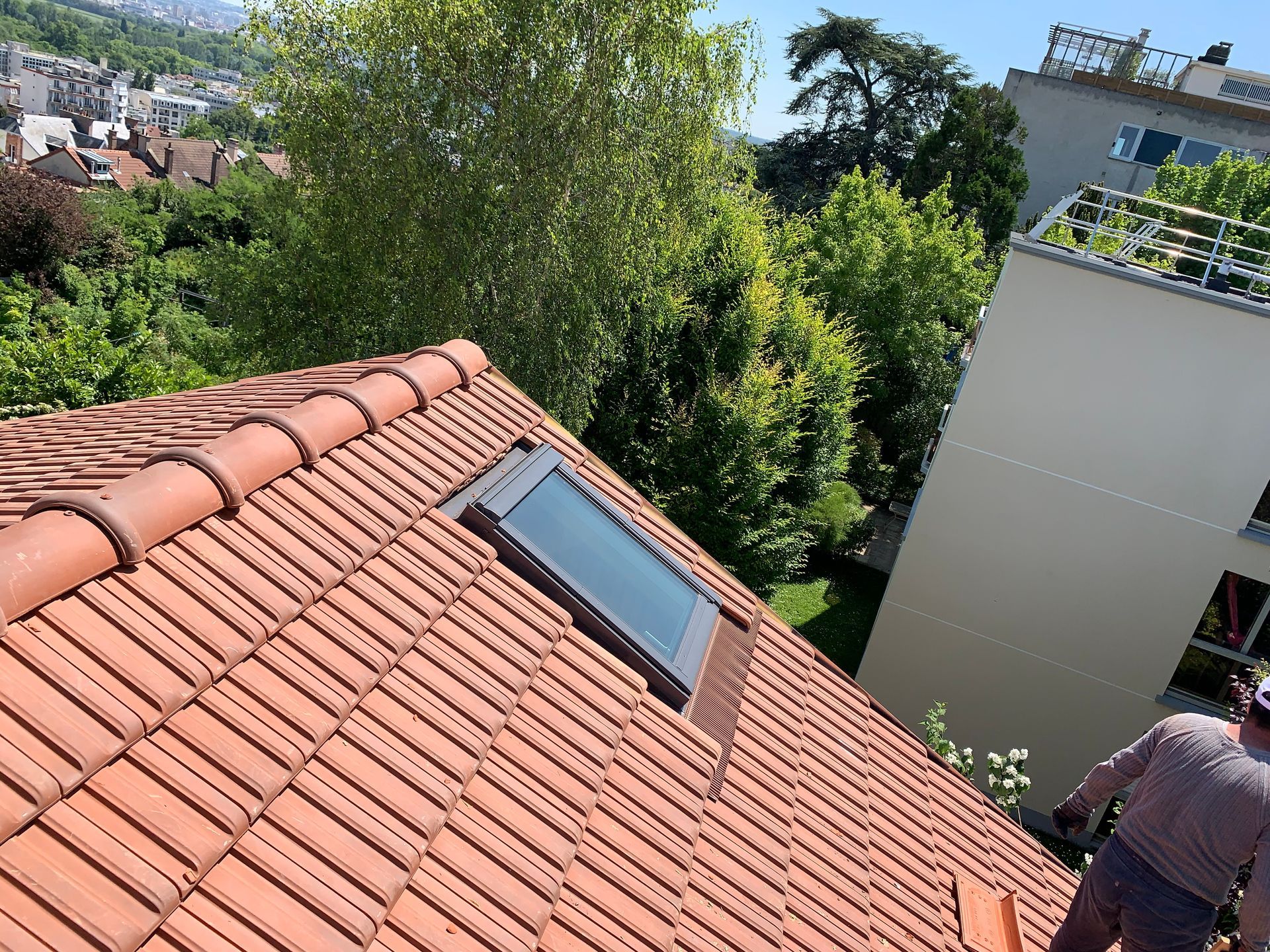 Toit de tuiles rouges avec puits de lumière à côté d'arbres feuillus et une personne au bord d'un bâtiment, surplombant la ville