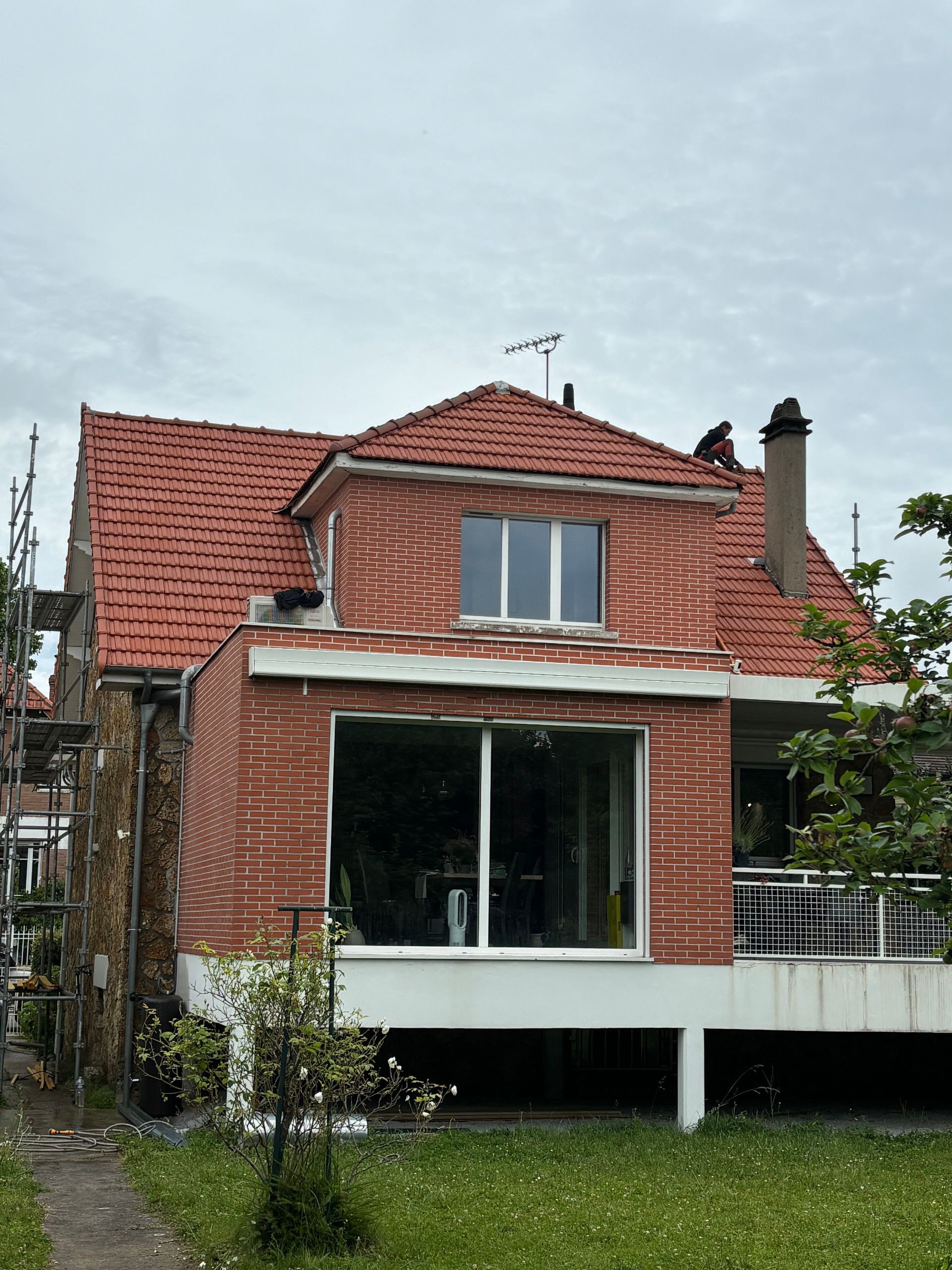 Maison en briques rouges avec un toit de tuiles rouges, de grandes fenêtres en façade et un porche surélevé sous un ciel nuageux