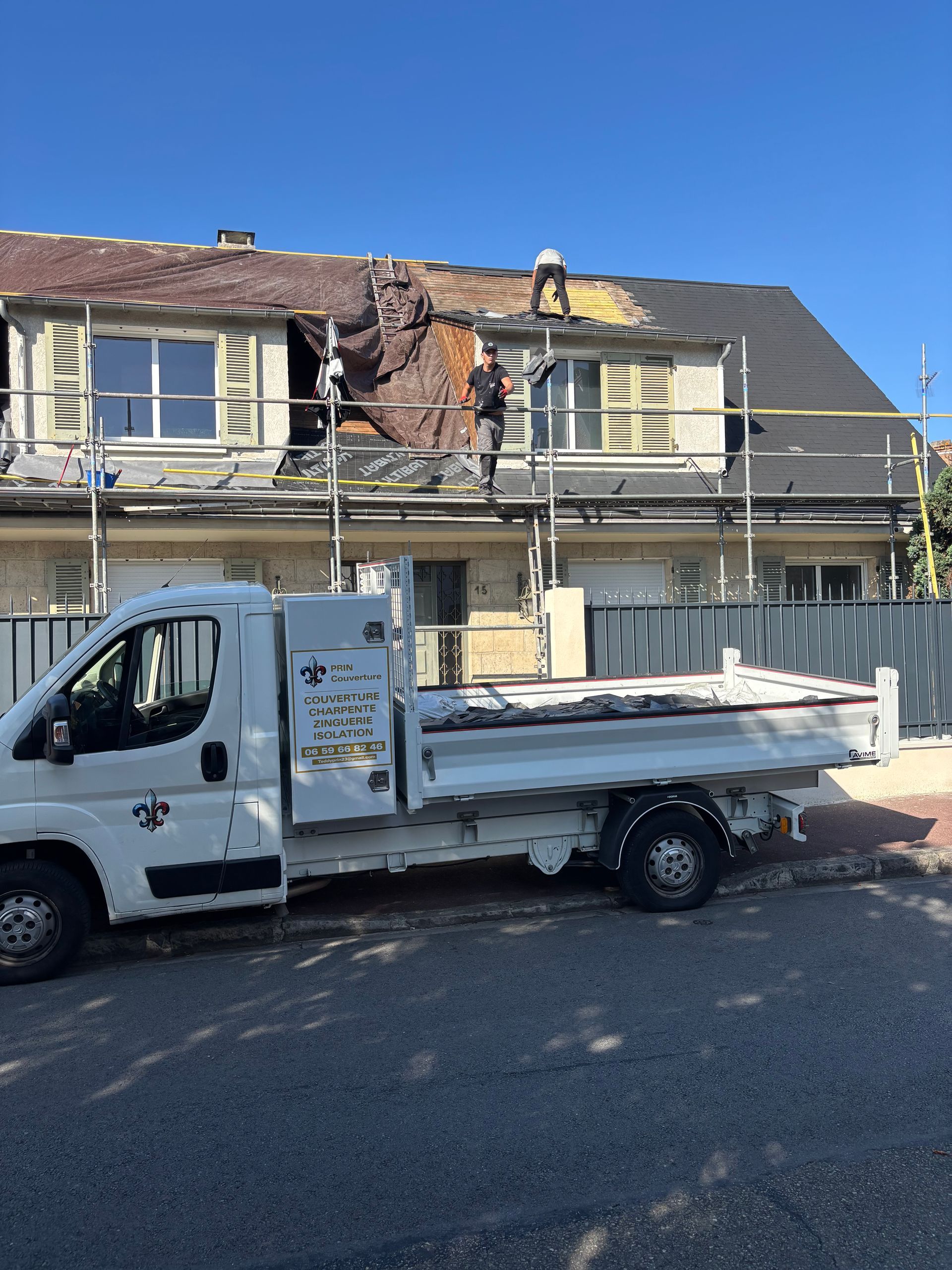 Un camion utilitaire blanc est garé devant une maison endommagée, au toit apparent et aux échafaudages.