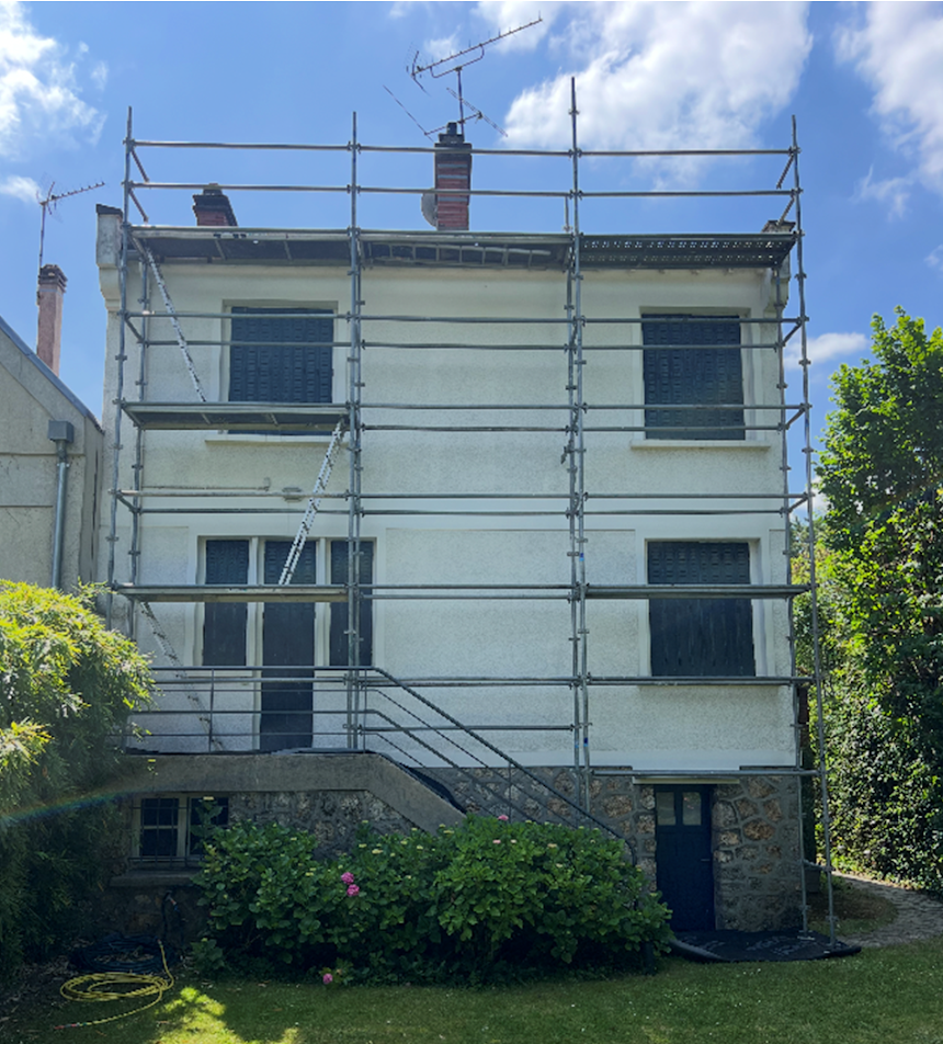 Maison blanche de trois étages avec des échafaudages en façade, sous un ciel bleu, avec un jardin devant.