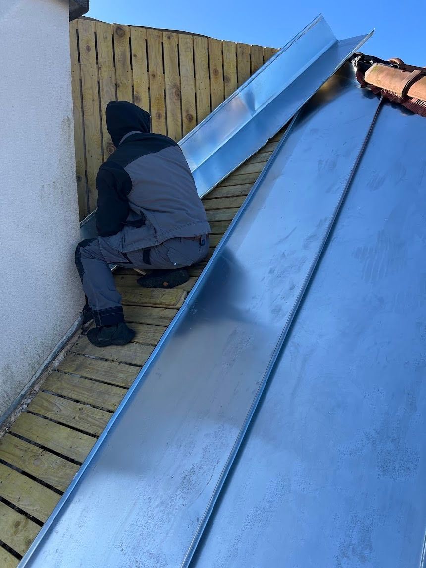 Un ouvrier accroupi sur un toit en pente, à côté d'un mur en bois et de panneaux de toiture en métal.
