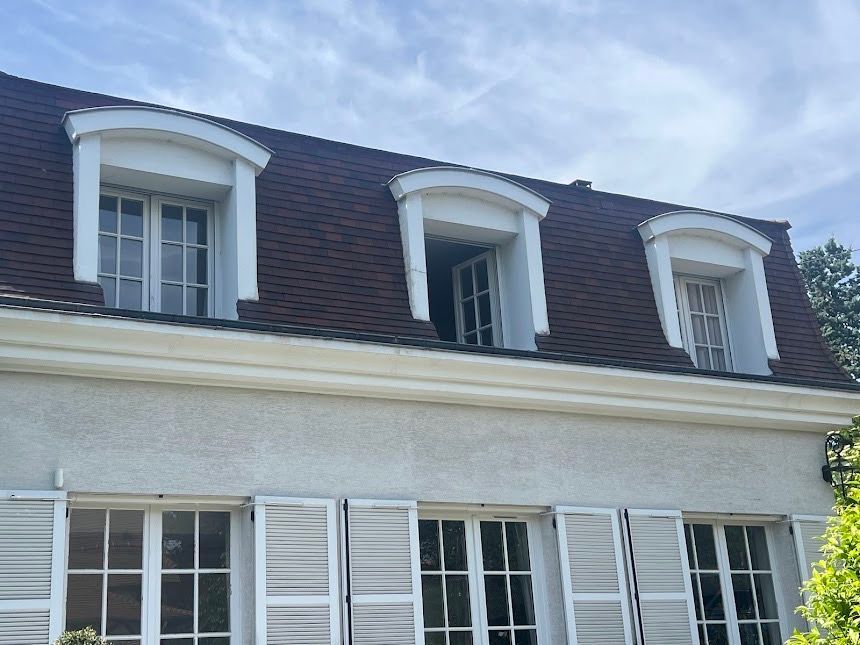Maison blanche avec toit en bardeaux foncés et lucarnes au-dessus des fenêtres à volets