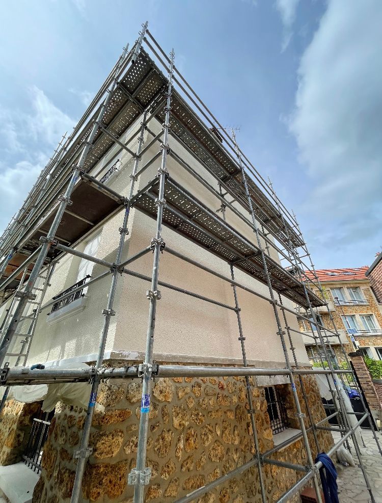Extérieur d'un bâtiment en rénovation, avec des échafaudages, des murs beiges et un soubassement en pierre partiellement recouvert.
