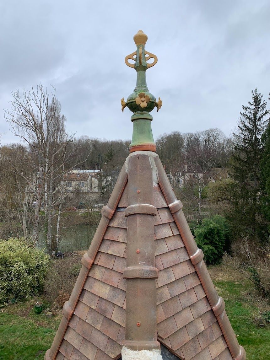 Fleuron de toit orné d'une croix dorée au sommet d'une tour carrelée, se détachant sur un ciel couvert.