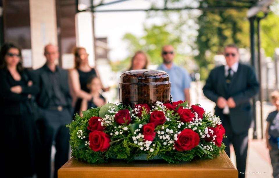 Ein mit roten und weißen Blumen bedeckter Sarg bei einer Beerdigung im Freien, im Hintergrund stehen Trauernde.