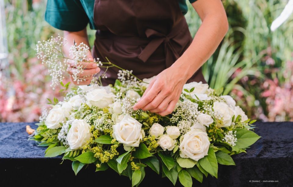 Hände arrangieren weiße Rosensträuße auf einem Tisch, zusammen mit Grünzeug und kleinen weißen Blüten.
