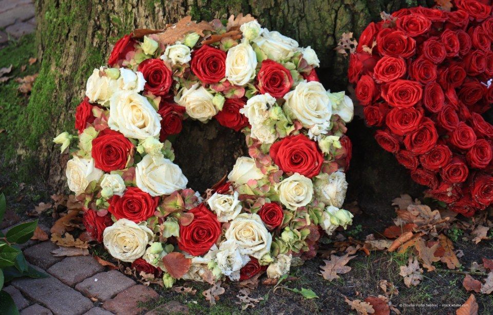 Blumenkränze aus roten und weißen Rosen an einem Baumstamm und einem Backsteinweg, daneben liegen herabgefallene Blätter.