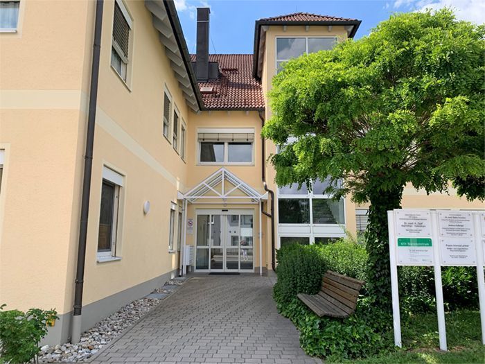 Das Gebäude mit seiner hellgelben Fassade, dem Eingang mit Glastüren und dem Ziegeldach unter strahlend blauem Himmel.