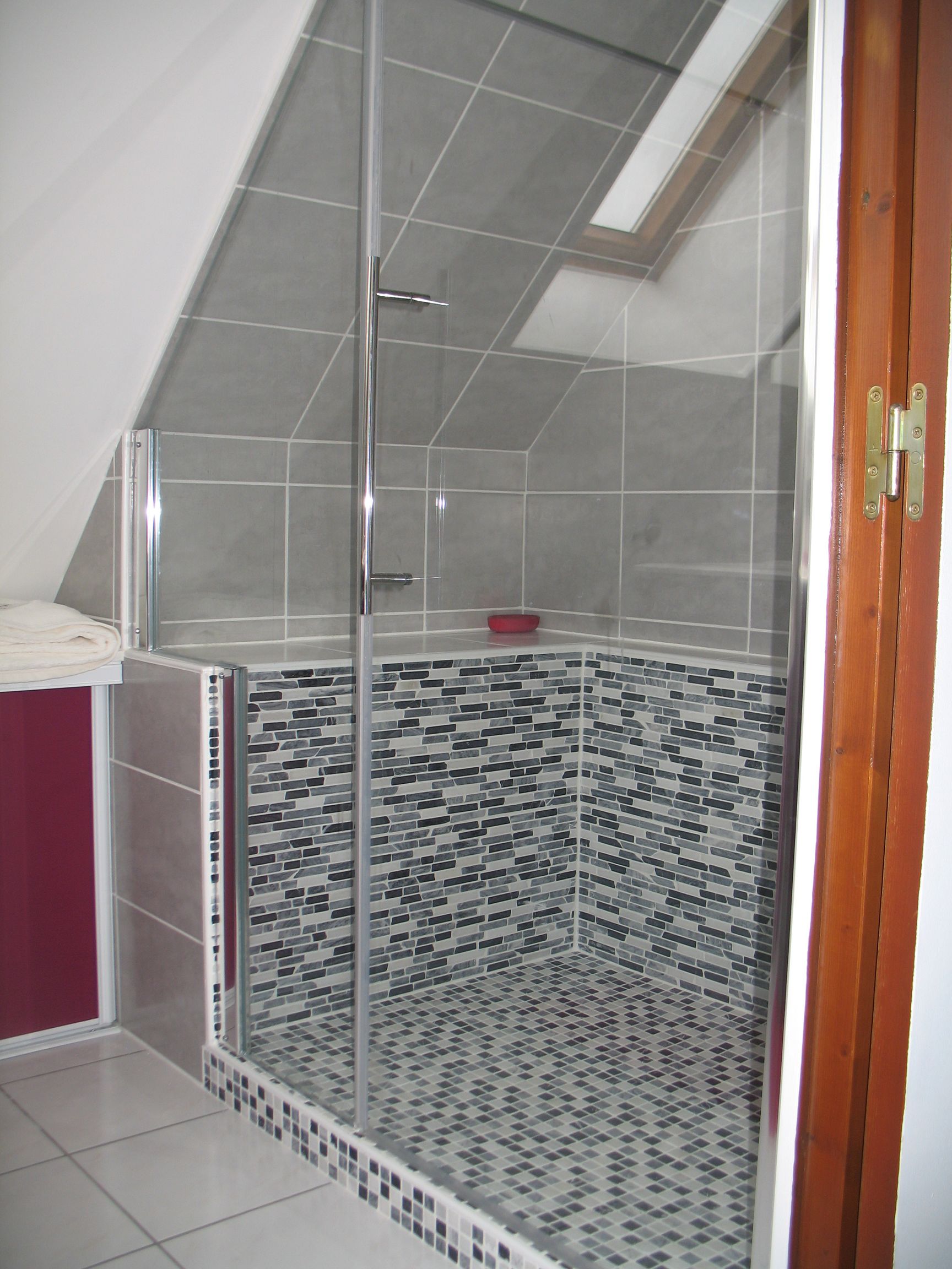 Douche avec portes vitrées et murs carrelés, mettant en valeur un mélange de mosaïque et de carreaux gris, sous un plafond incliné.