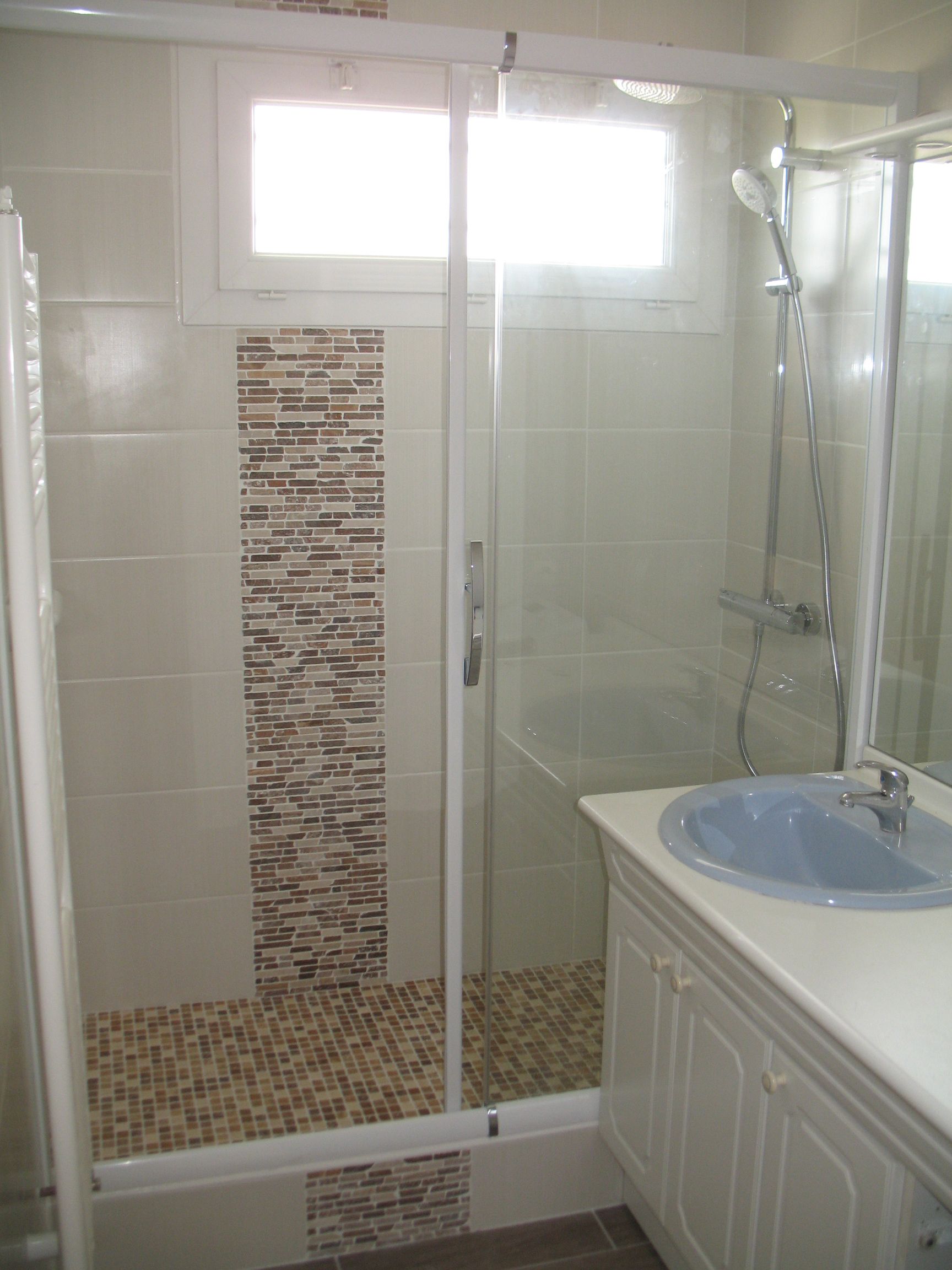 Salle de bain avec douche en verre, lavabo bleu, carrelage à motifs et fenêtre.