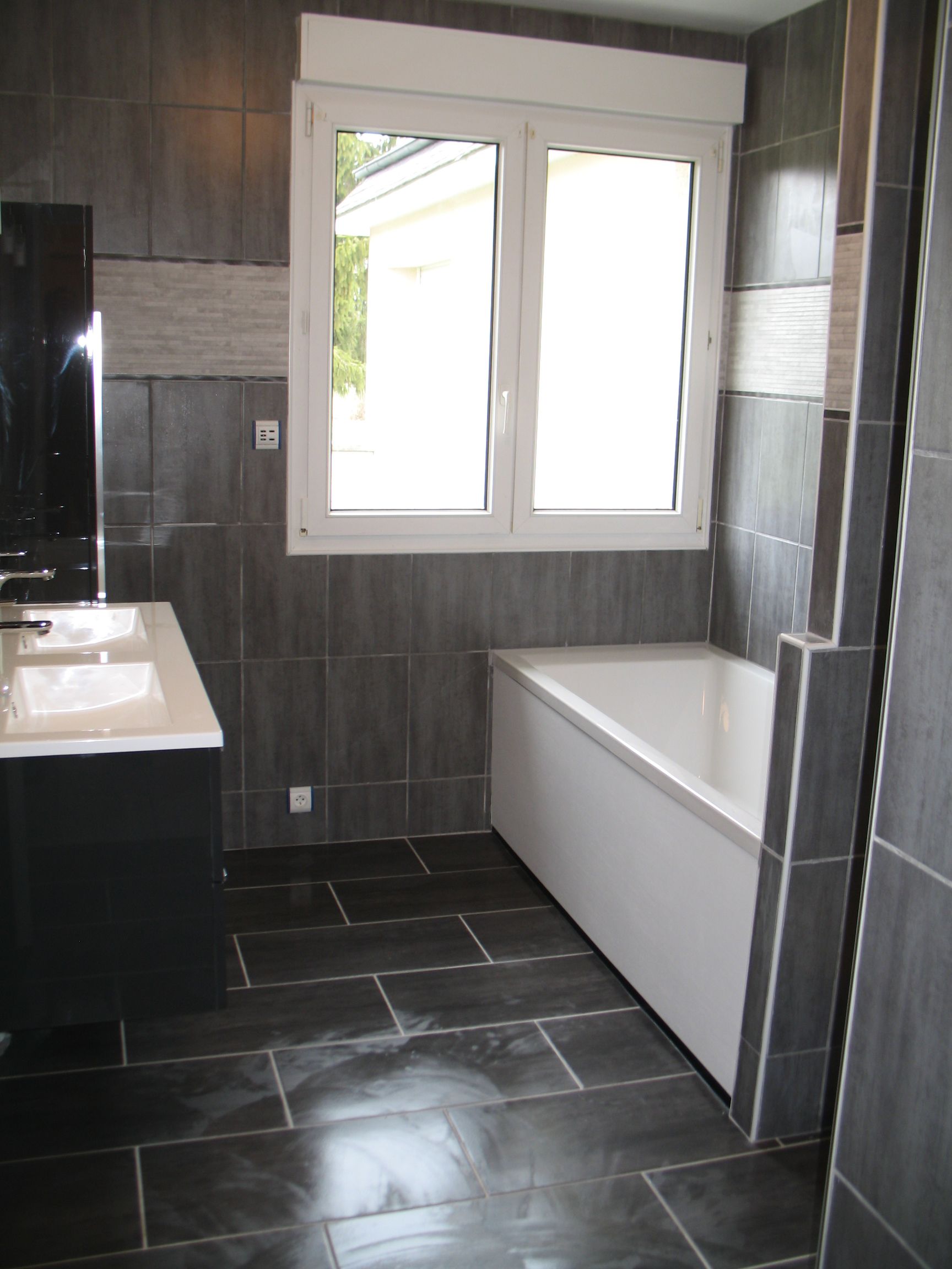 Salle de bain avec carrelage foncé au sol et aux murs, baignoire blanche et fenêtre.
