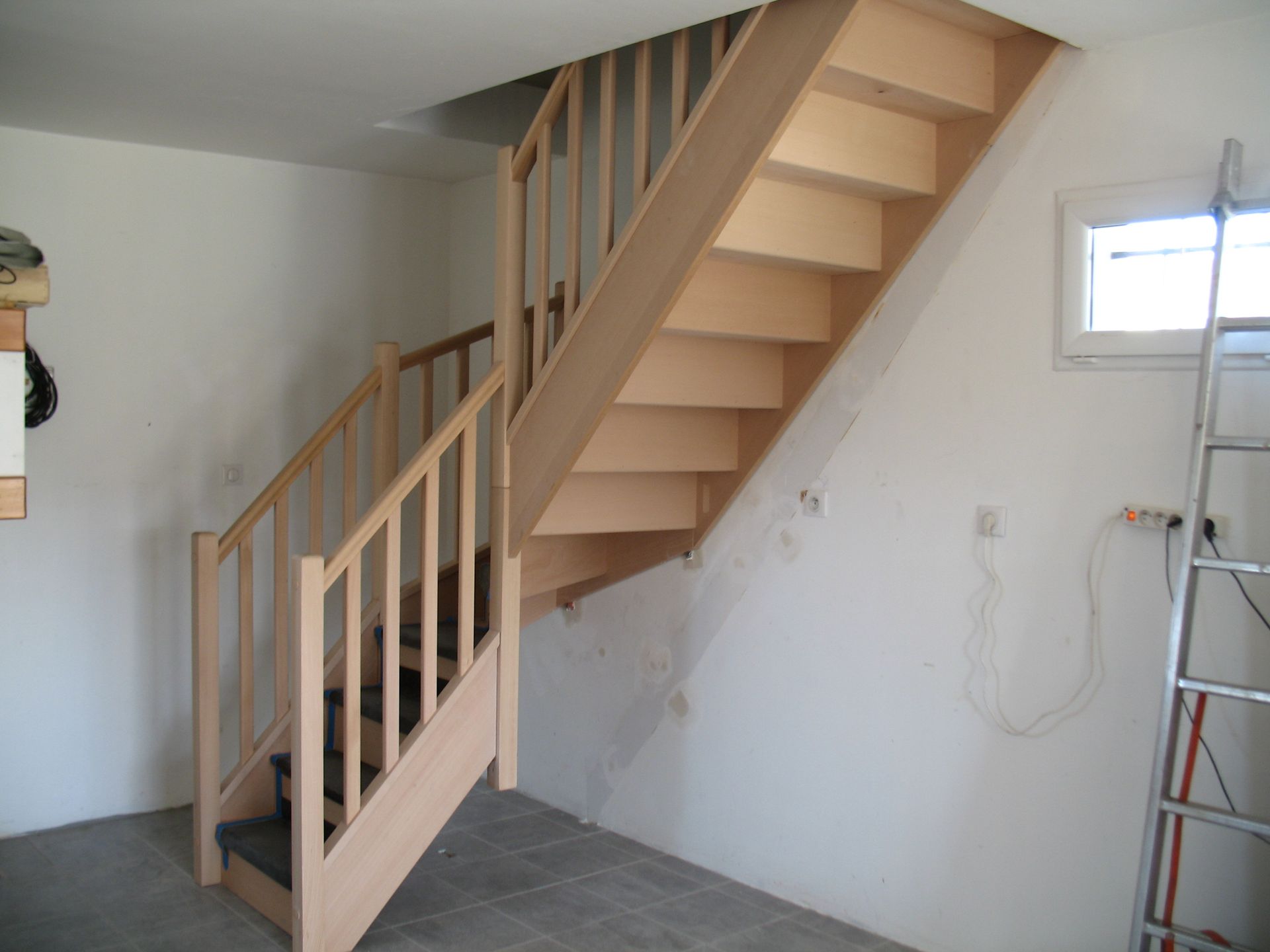 Escalier en bois avec rampe menant à un niveau supérieur, situé dans une pièce aux murs blancs avec une fenêtre et une échelle.