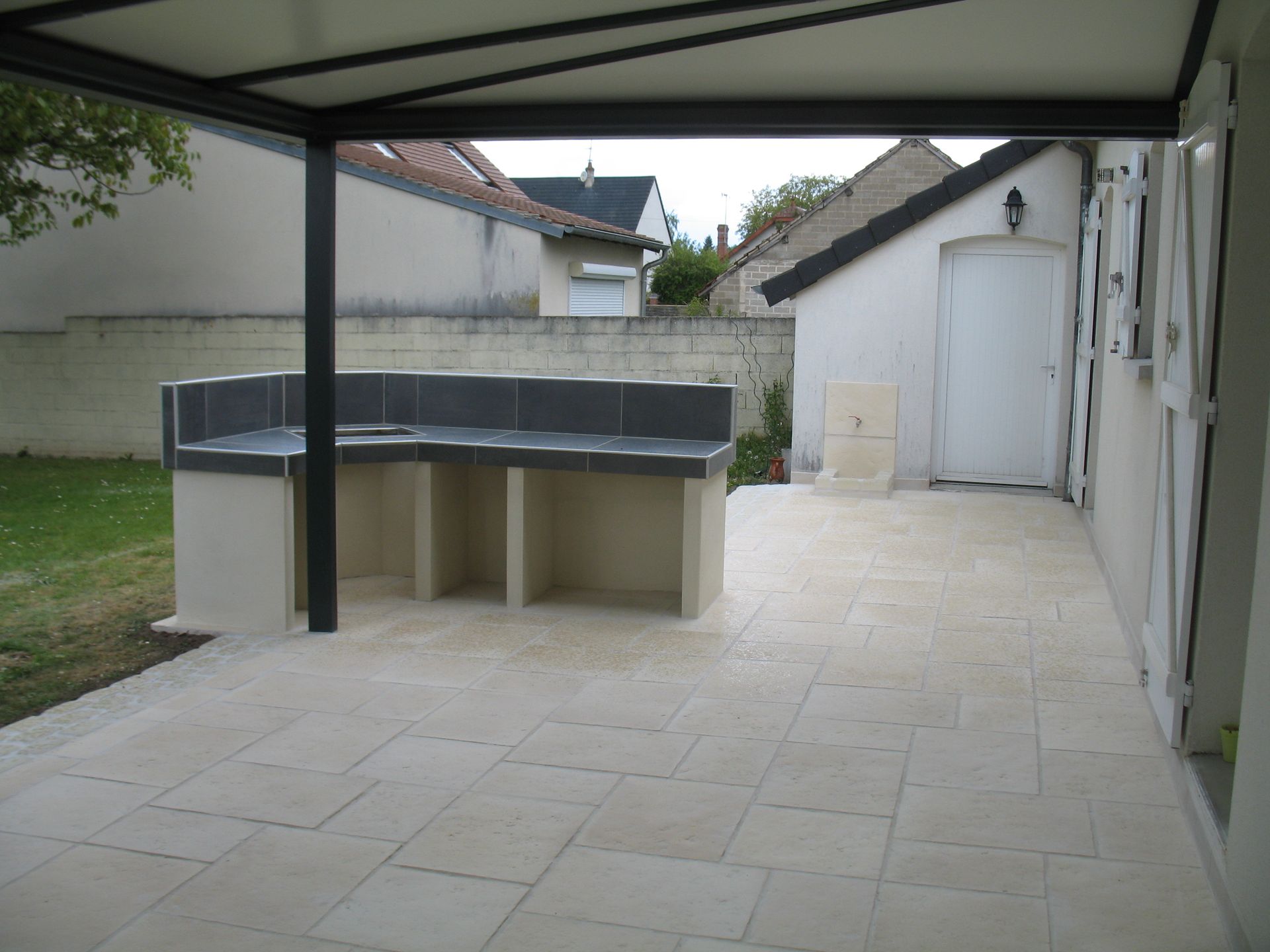 Terrasse extérieure avec comptoir intégré et pergola noire. Carrelage crème, murs blancs et pelouse.