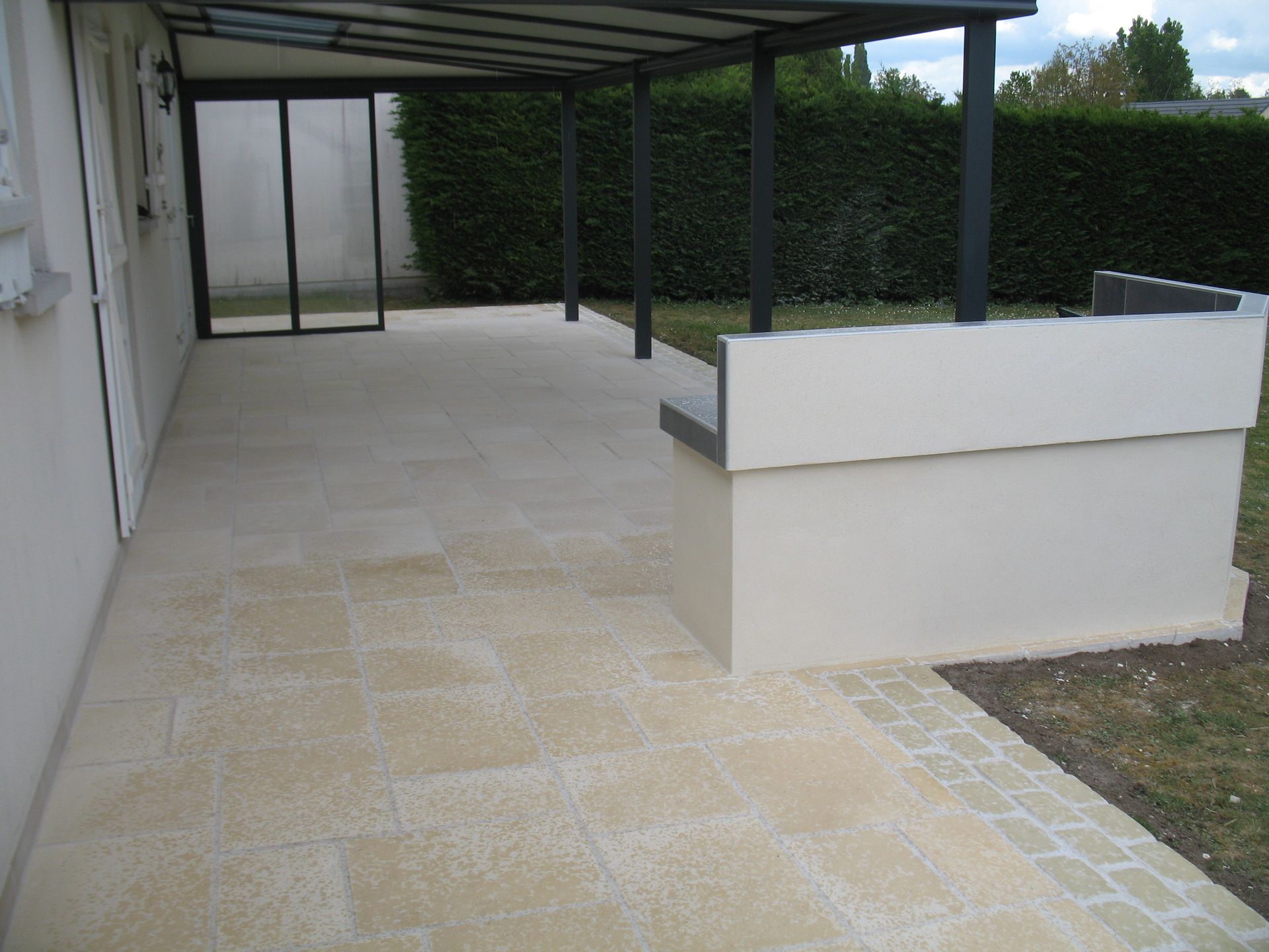 Terrasse extérieure pavée beige, attenante à une maison couleur crème. Auvent à cadre noir et mur blanc à droite.