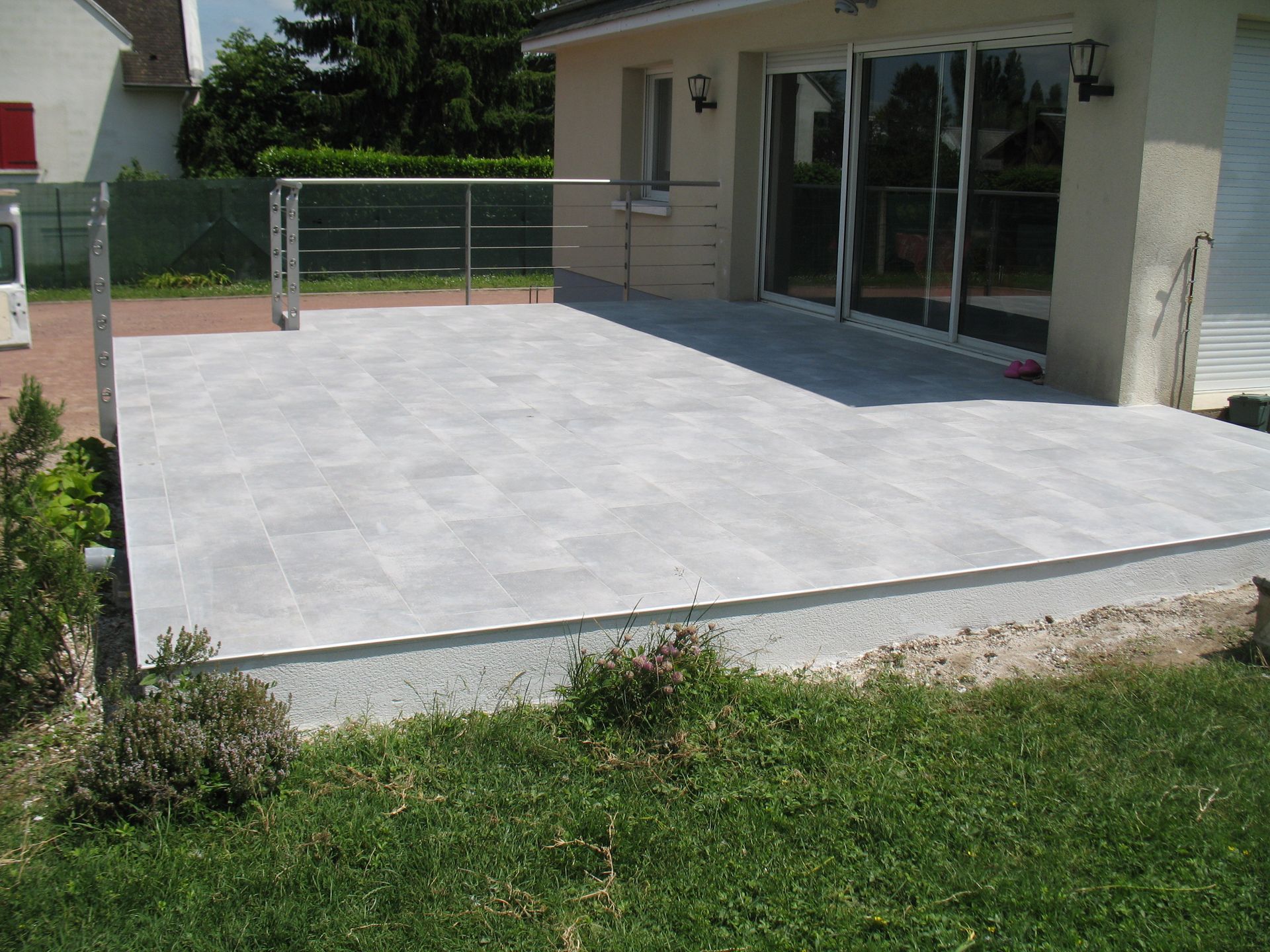 Terrasse carrelée grise à l'extérieur d'une maison avec portes vitrées, balustrade et herbe.