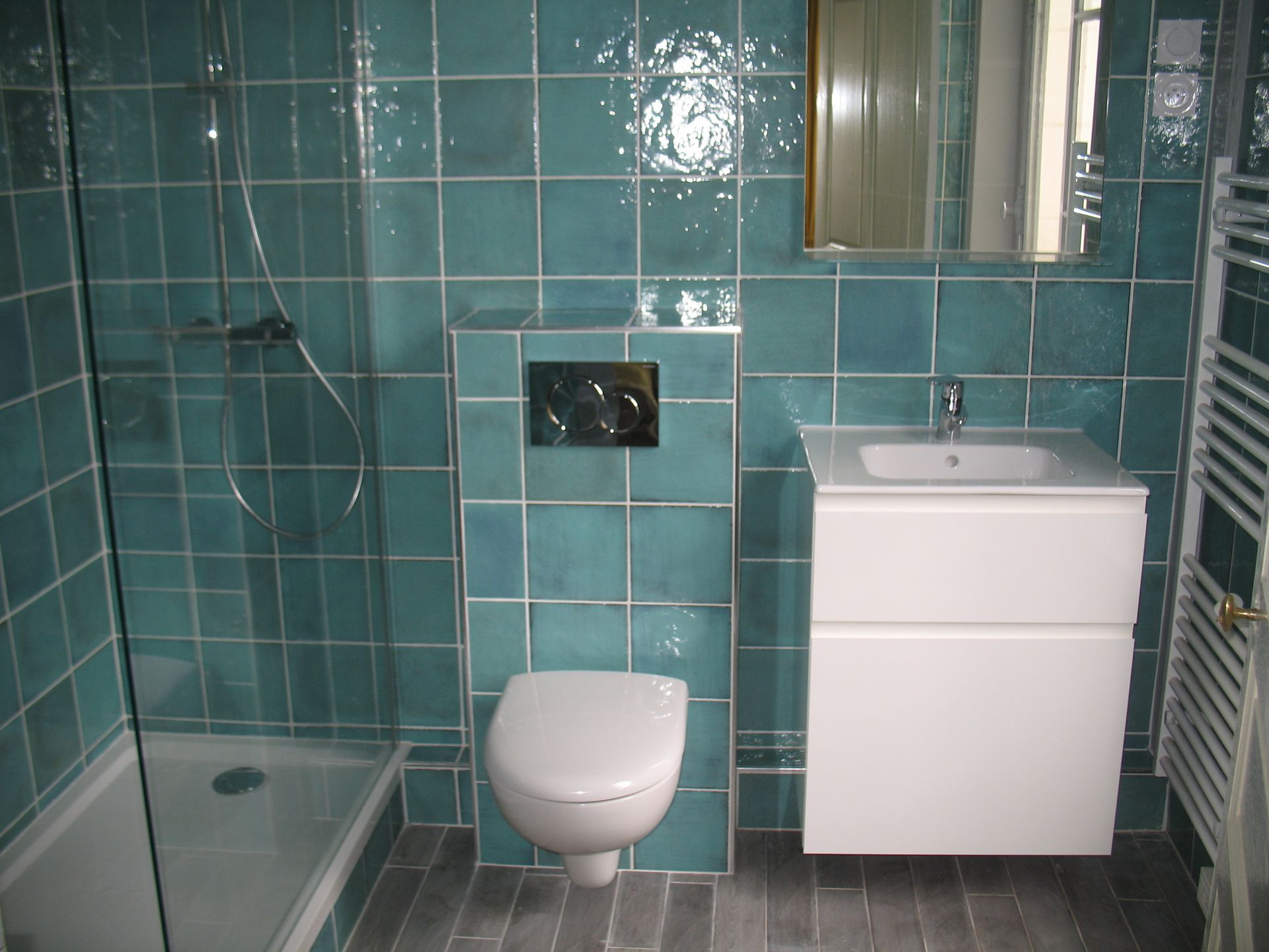 Salle de bains carrelée turquoise avec douche, toilettes, lavabo et miroir. Sol gris.