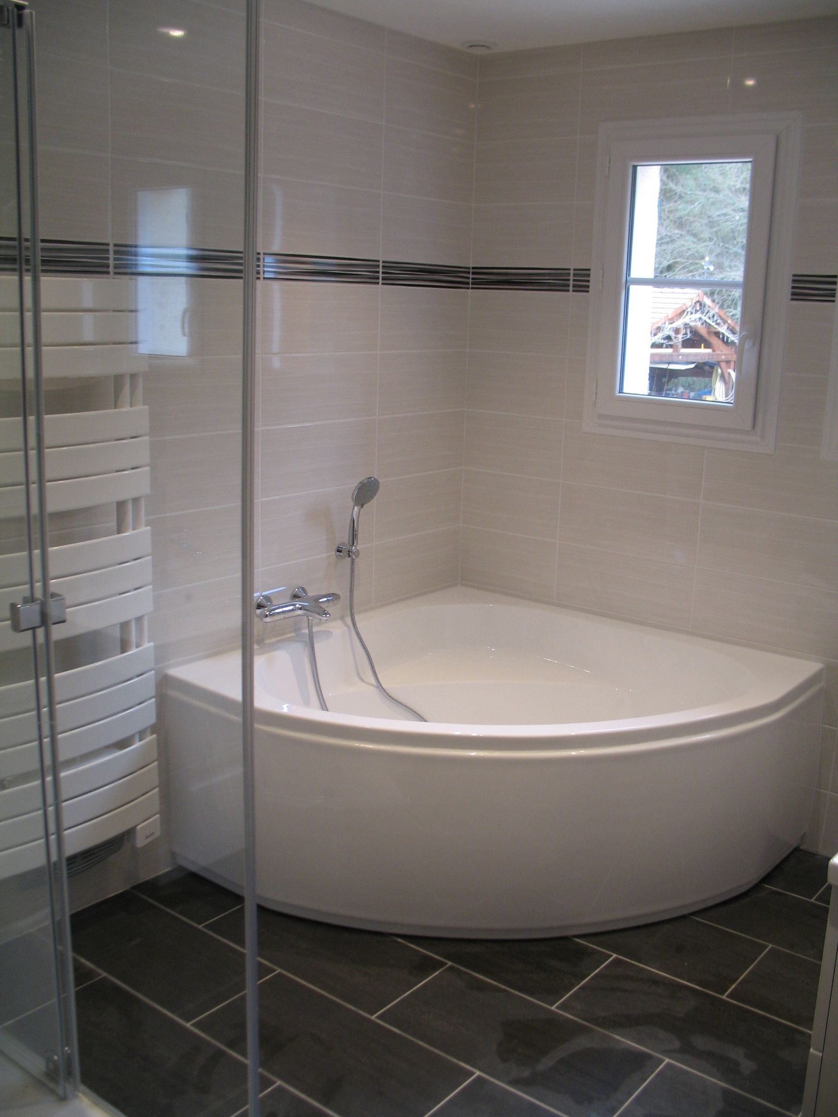 Baignoire d'angle dans une salle de bain carrelée, avec une petite fenêtre et un porte-serviettes, sol noir et une douche.