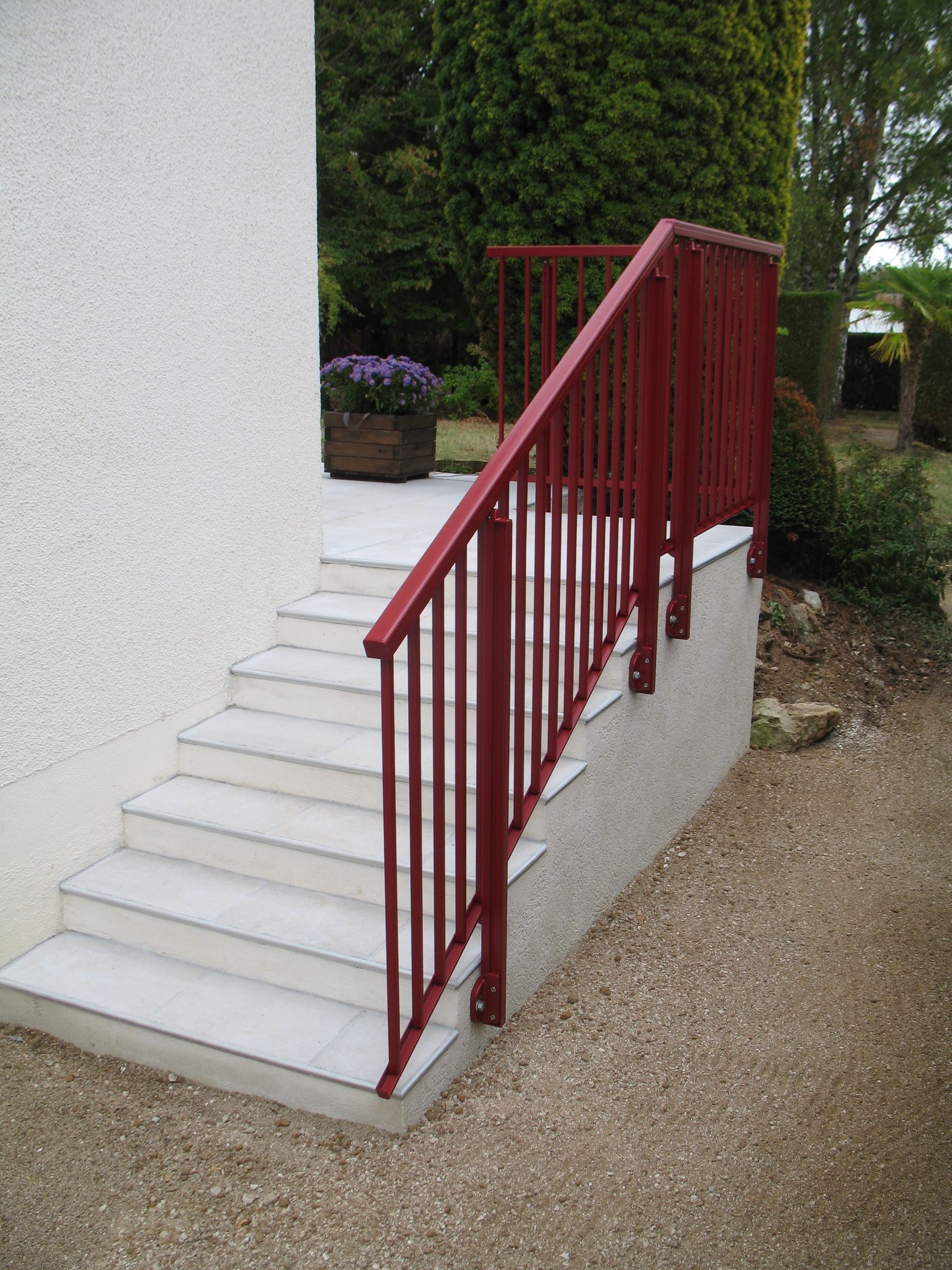 Rampe rouge sur un escalier extérieur en béton à côté d'un mur en stuc blanc ; fleurs violettes en arrière-plan.