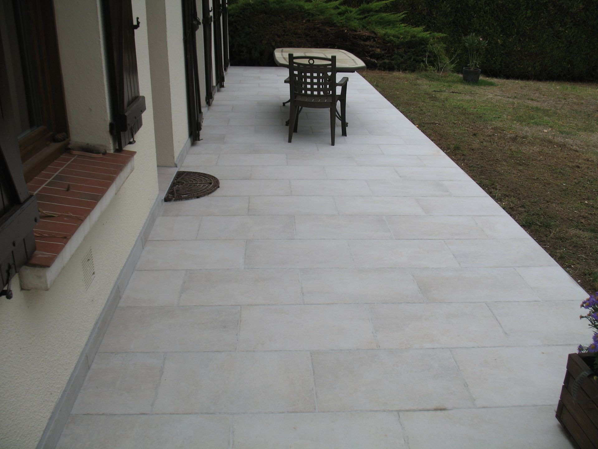 Patio en pierre claire à côté d'une maison aux volets marron. Une table et une chaise y sont installées ; l'herbe en arrière-plan.