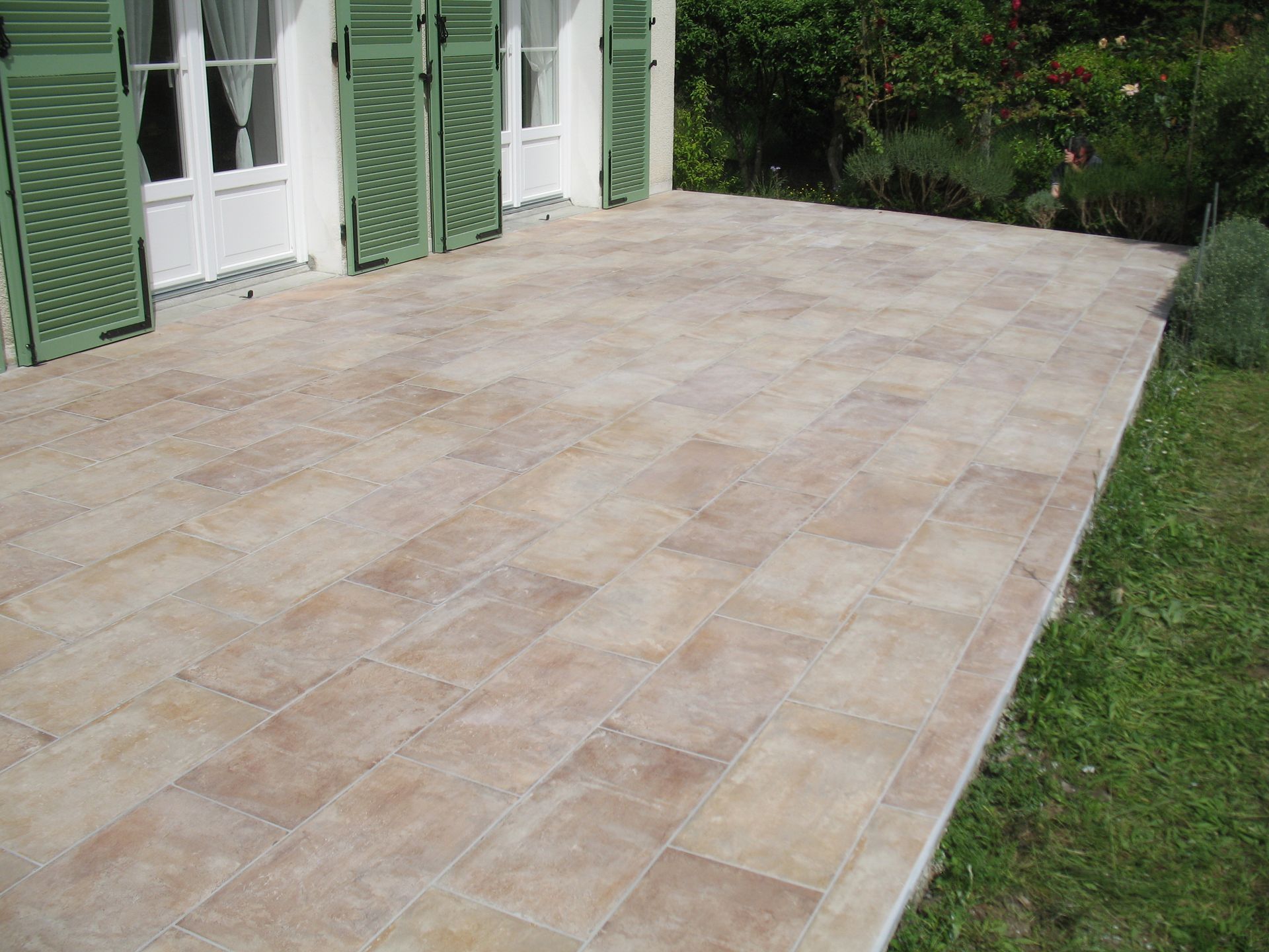 Un patio aux carreaux beiges devant un bâtiment blanc aux volets verts, bordé d'herbe verte.