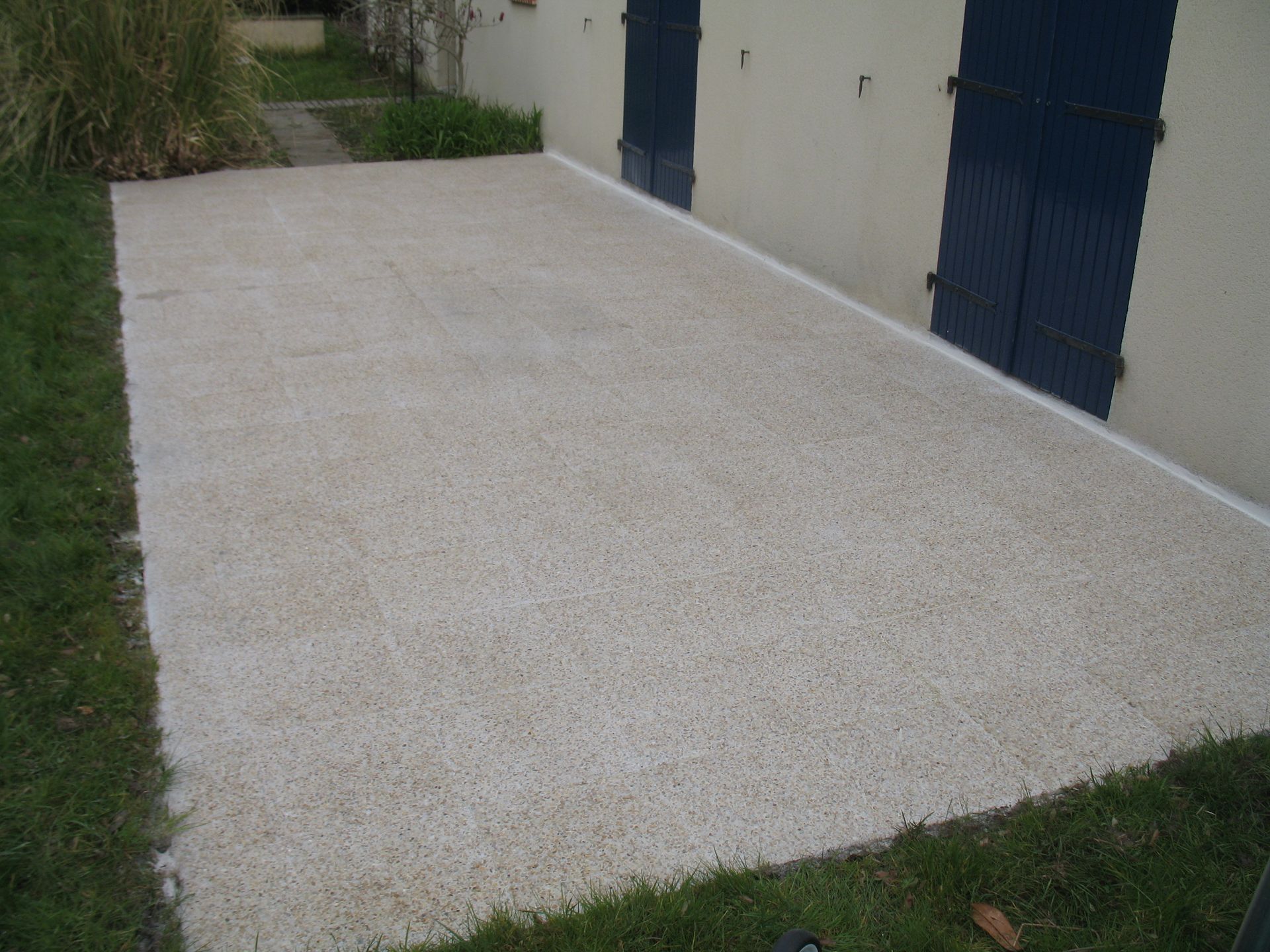 Terrasse en béton à côté d'un immeuble aux volets bleus, entourée de gazon.