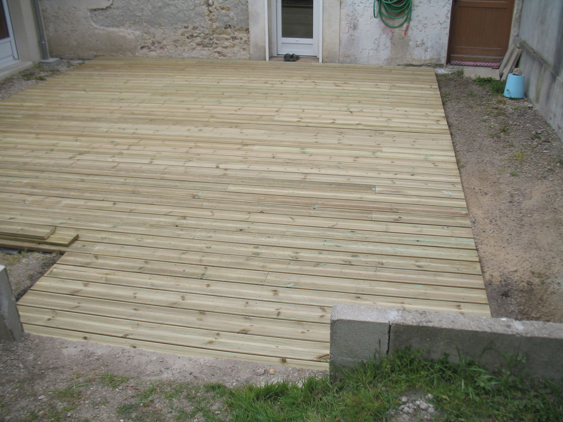 Terrasse en bois construite contre un bâtiment avec une porte. La terre et l'herbe entourent la terrasse.